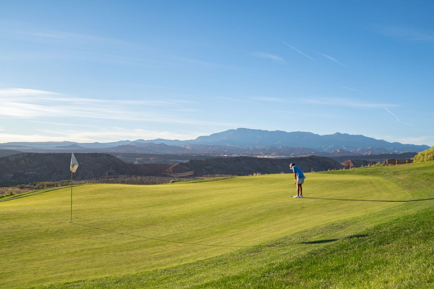 Golfer on lush green golf course fairway with mountain range and blue sky backdrop on a sunny day.