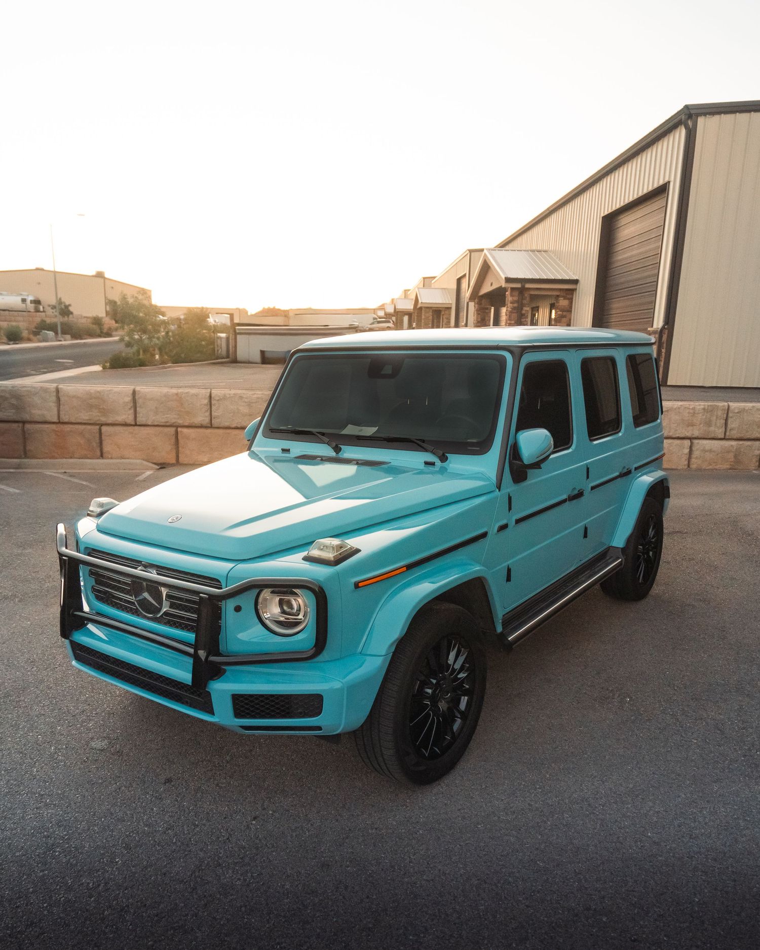 A teal Mercedes-Benz G-Wagon SUV parked in a desert setting with industrial buildings in the background at sunset.