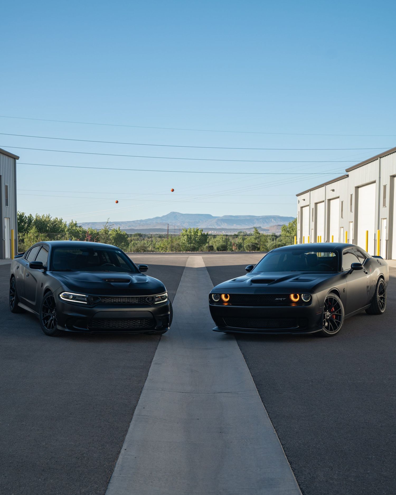Two matte black Dodge muscle cars, a Charger and Challenger, parked facing each other outside storage units.