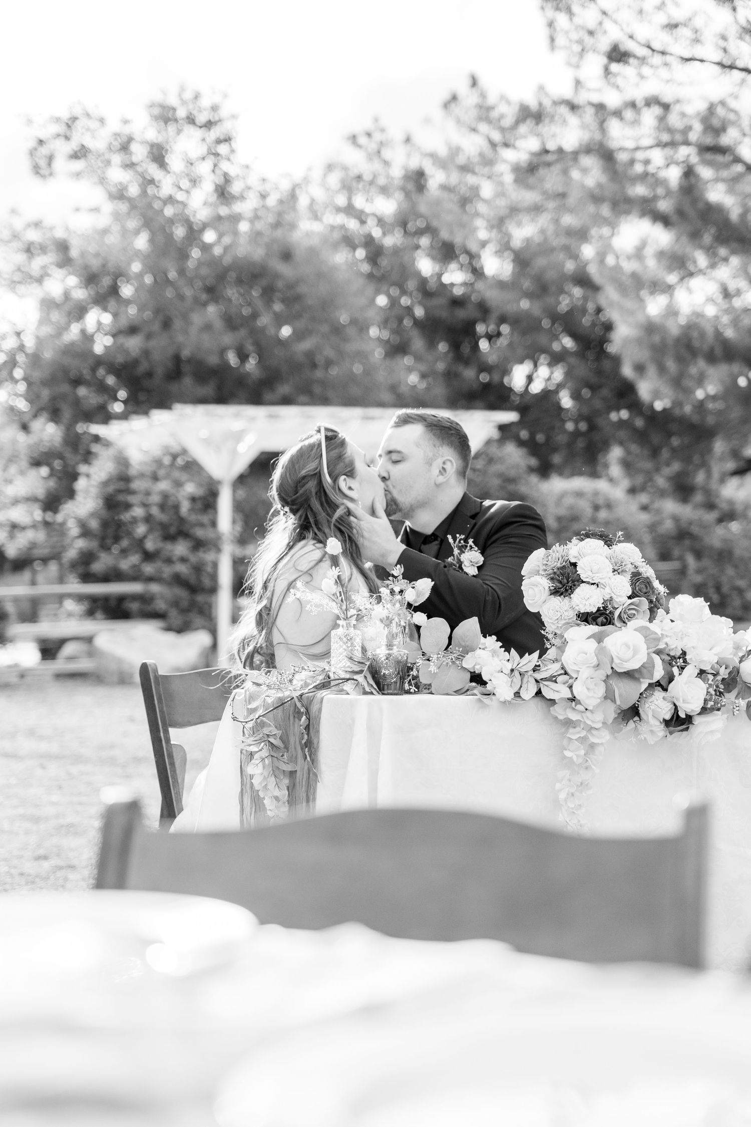 Black and white photo of a bride and groom sharing a kiss at their sweetheart table during an outdoor wedding reception.