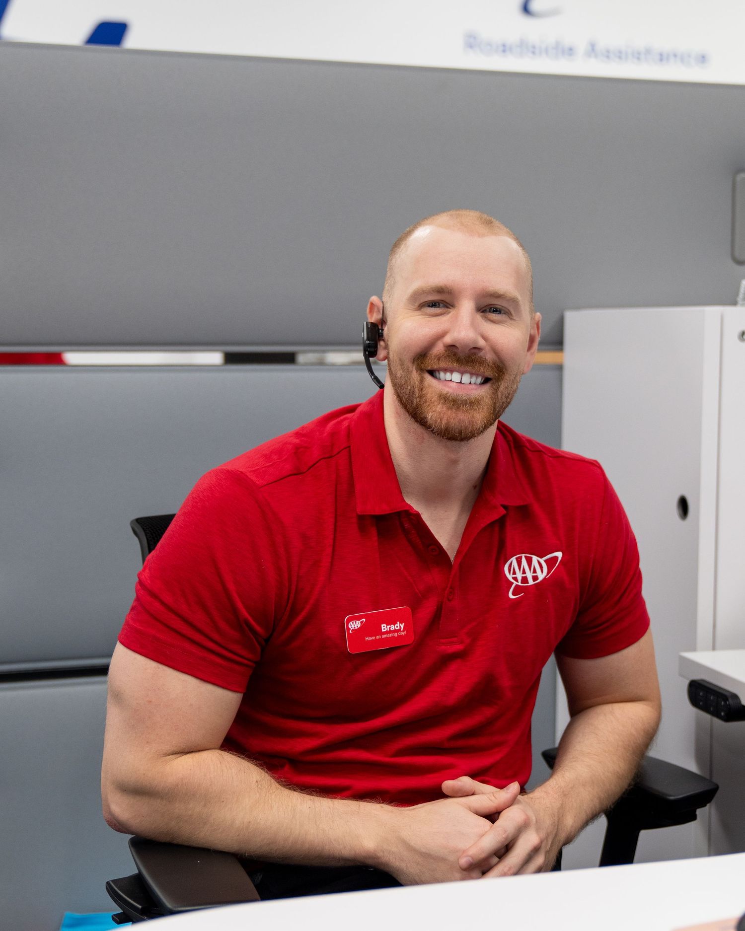 Smiling AAA customer service representative wearing a red polo shirt and headset seated at a desk in an office.