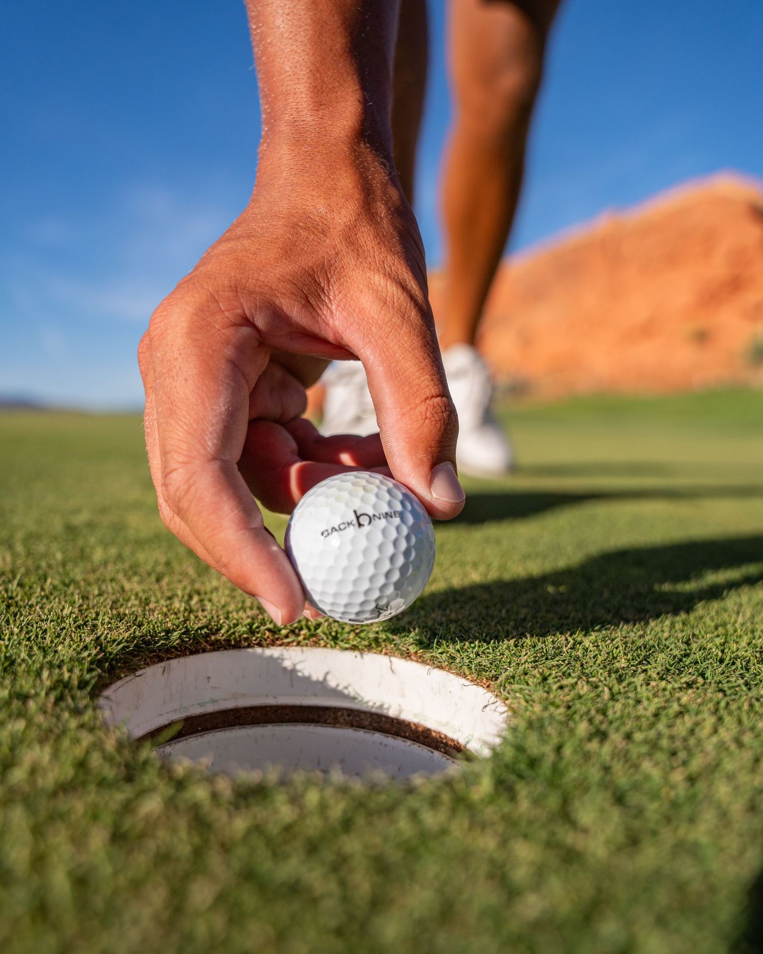 Golfer retrieving a white golf ball from the cup on a sunny green course with red rock formations in the background.