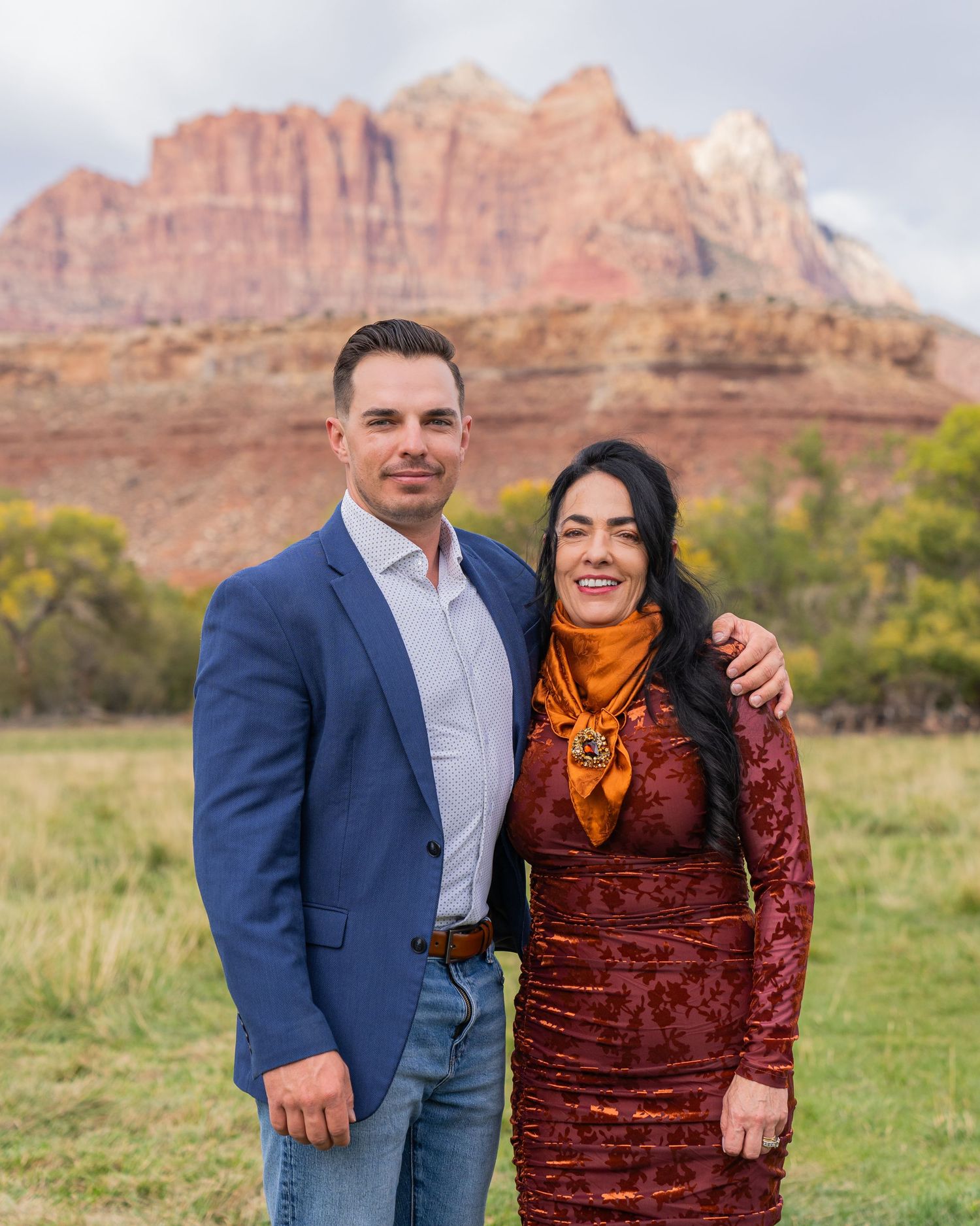 Couple posing together in front of red rock canyon landscape, man in blue blazer, woman in burgundy dress.