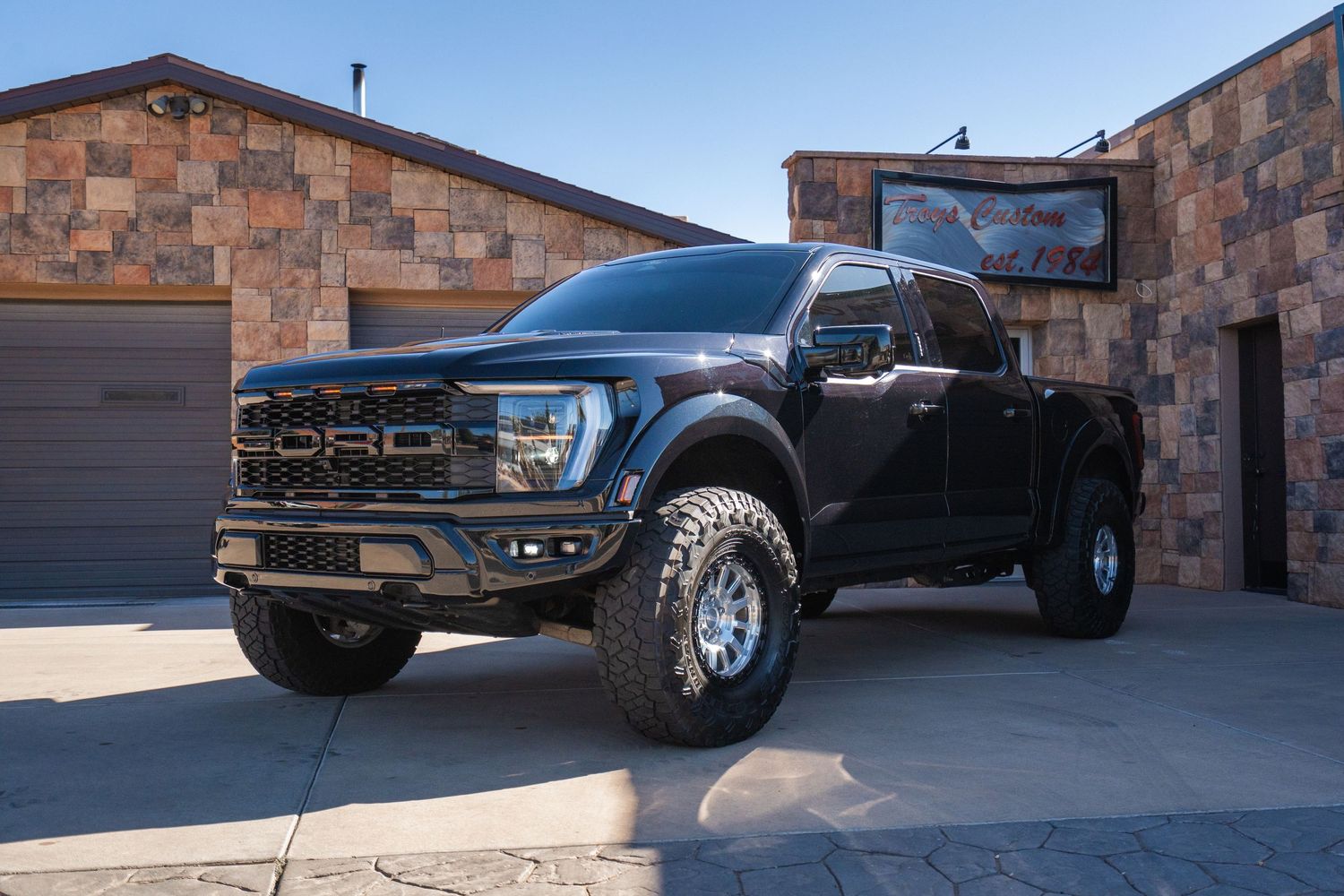 Black lifted Ford F-150 Raptor truck parked outside a stone building with garage doors on a sunny day.