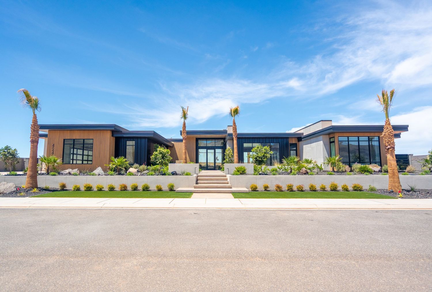Modern single-story home with dark trim, large windows, palm trees, and lush landscaping under a bright blue sky.