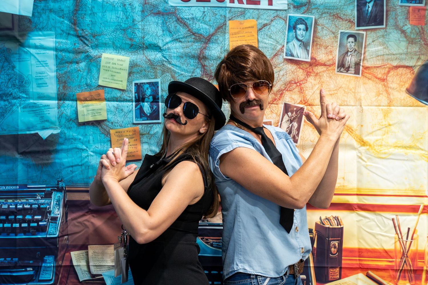 Two people wearing fake mustaches and hats pose back-to-back in front of a colorful spy-themed backdrop.
