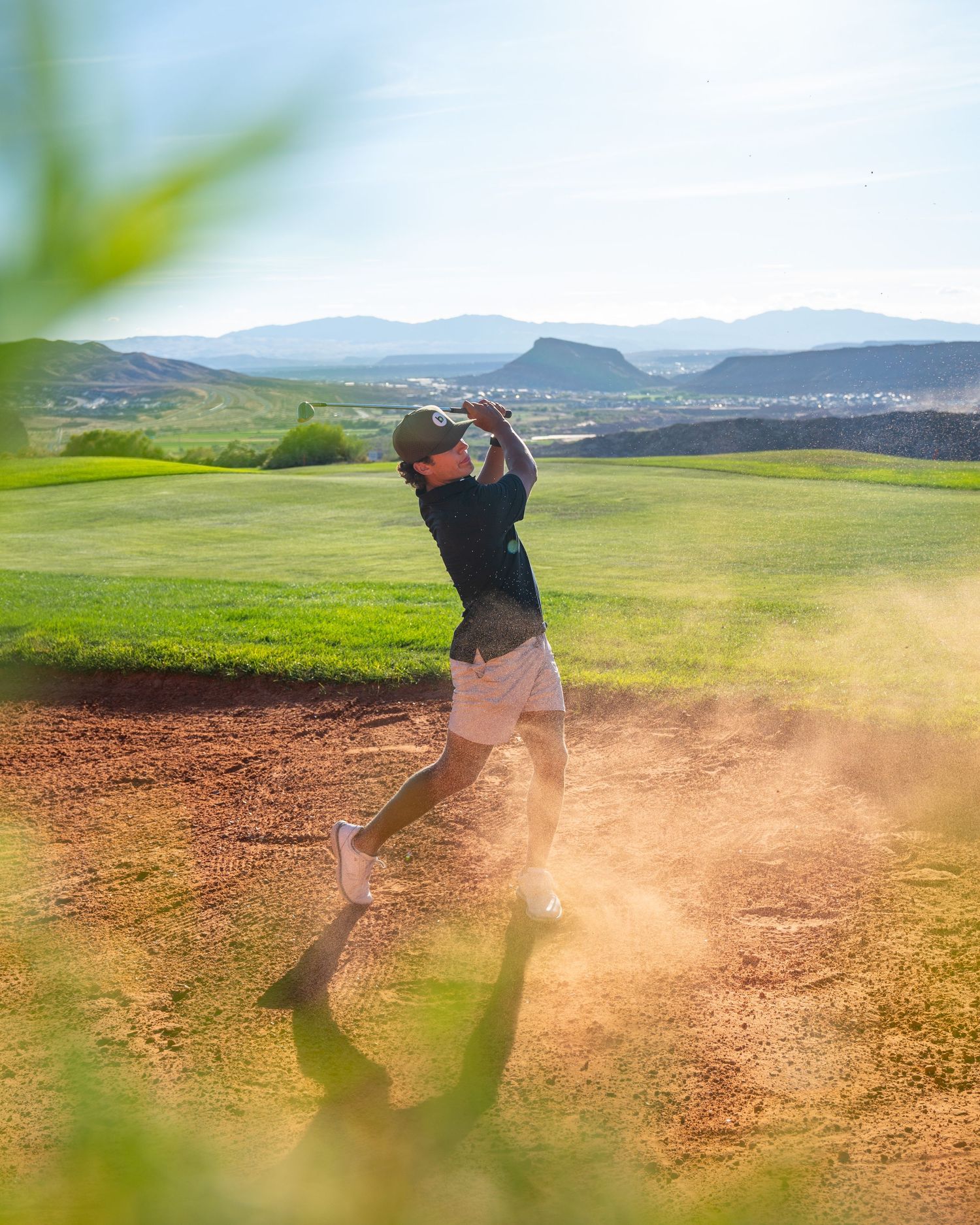 Golfer swinging club from sandy bunker on lush green course with mountain backdrop in bright sunlight.