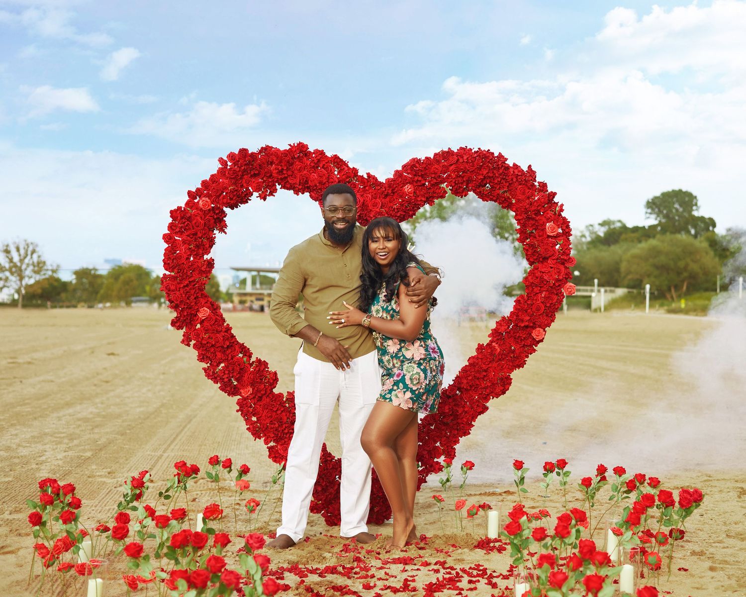 Large heart shapes made from red rose petals on sandy beach with blue sky.