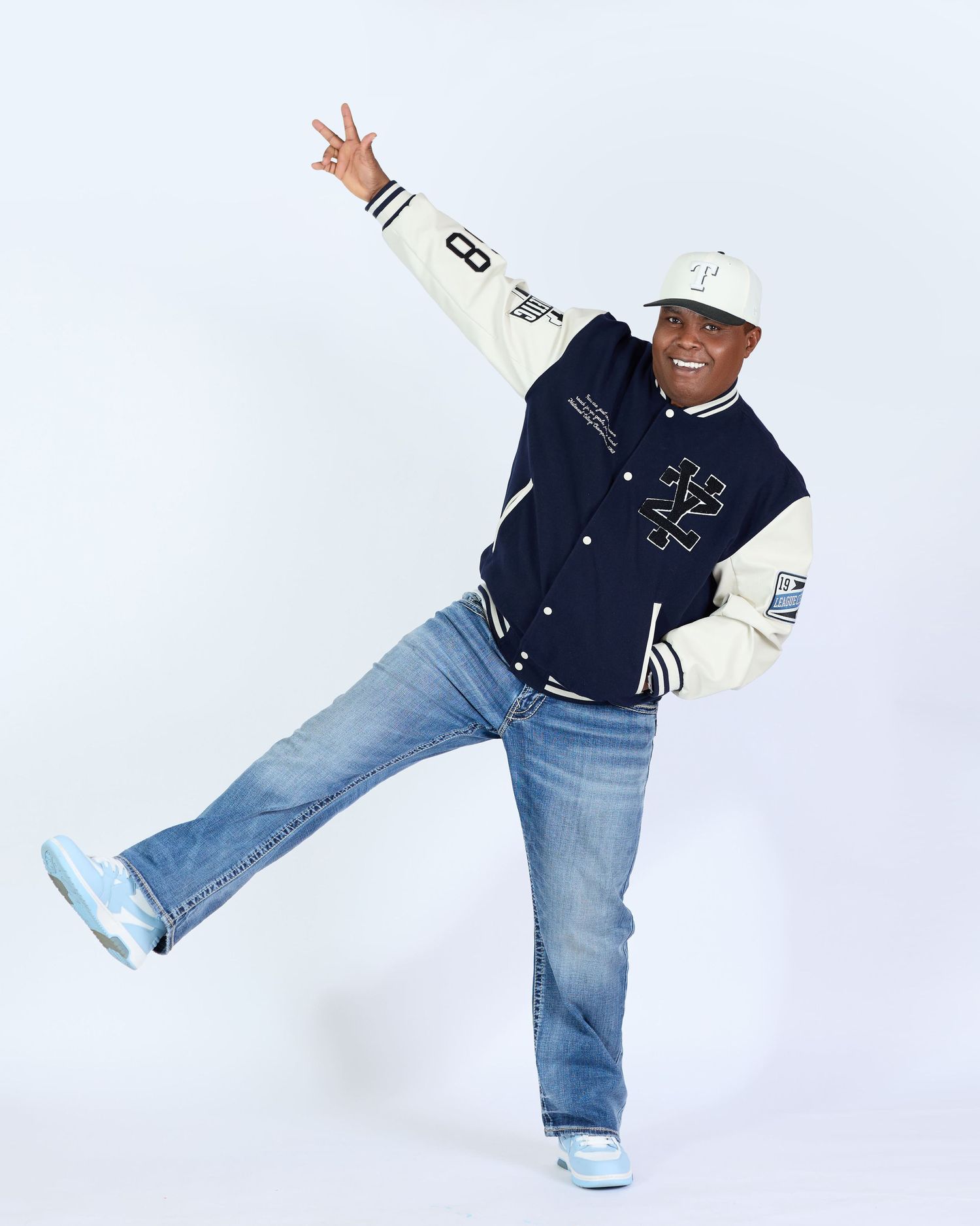 Playful jumping pose in varsity jacket and jeans against white background.
