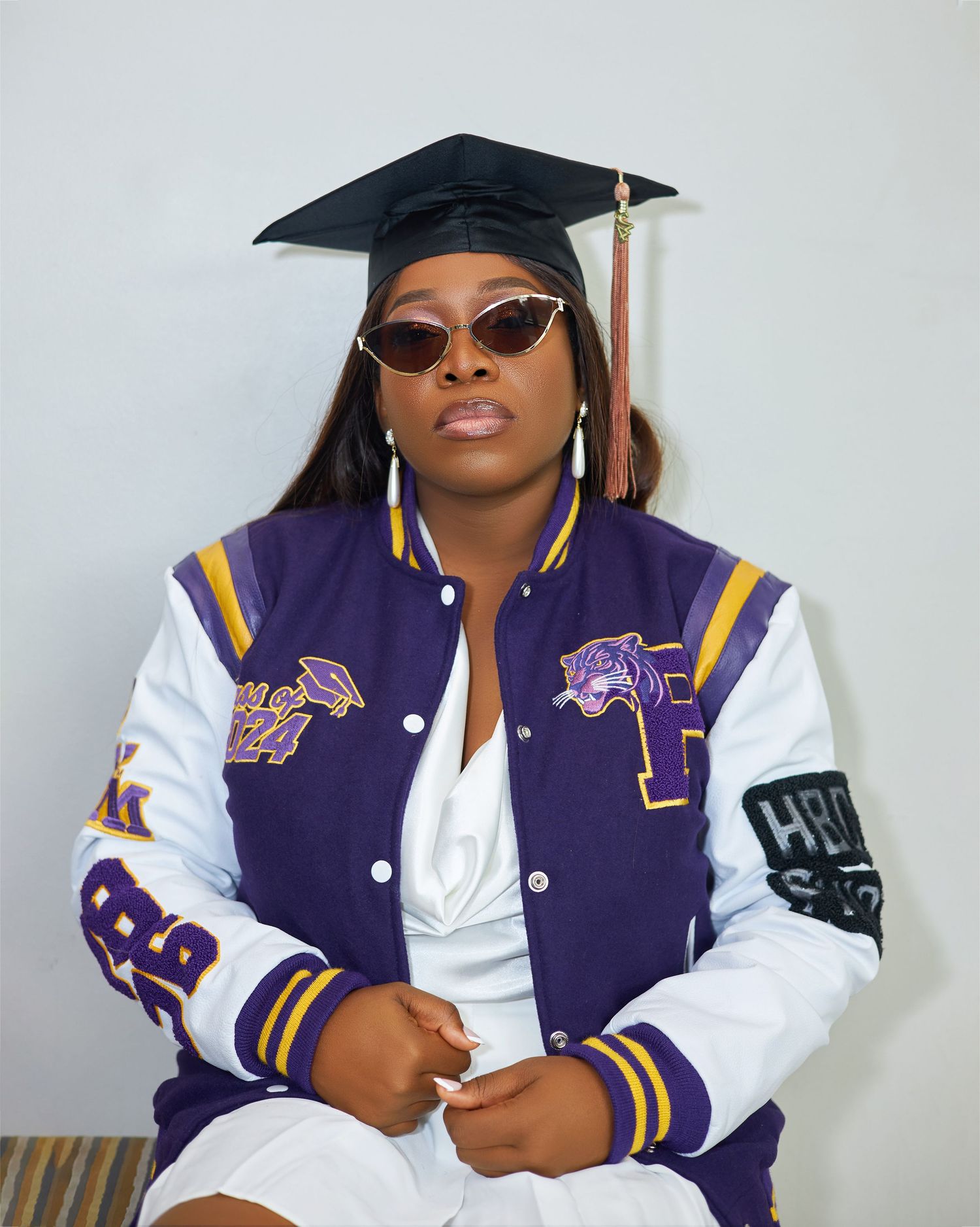 Graduate wearing purple and white varsity jacket poses with sunglasses and diploma for graduation photos.