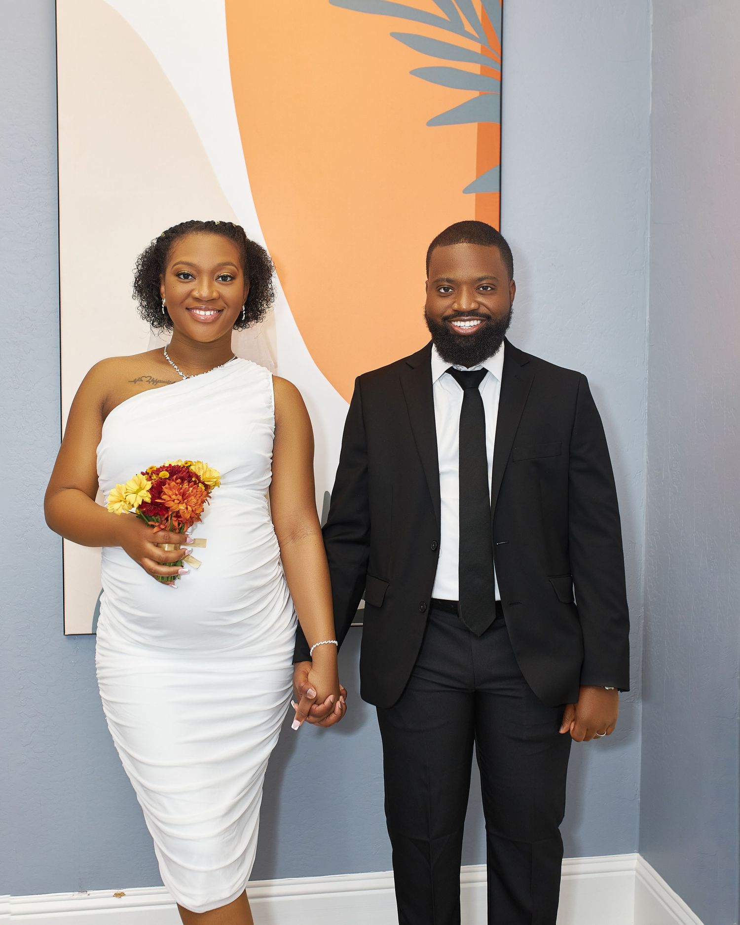 Wedding couple poses against grey wall in white dress and black suit.