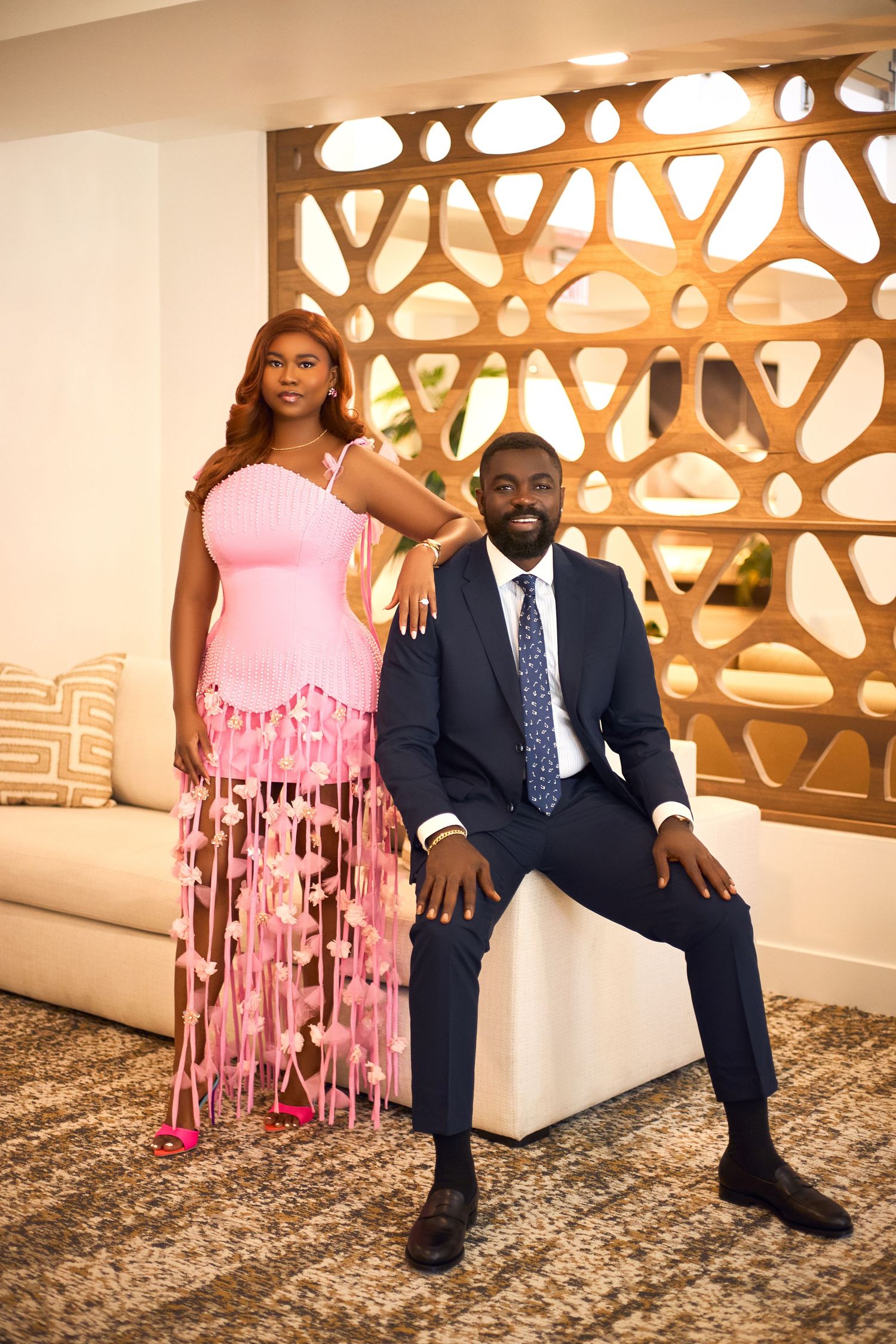 Couple poses elegantly in formal attire against decorative wall with pink flowy dress.
