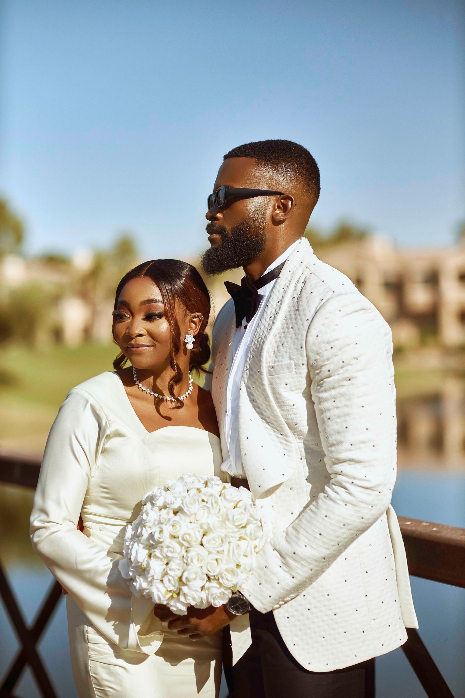 A couple in white attire poses with a bouquet near a waterfront setting.