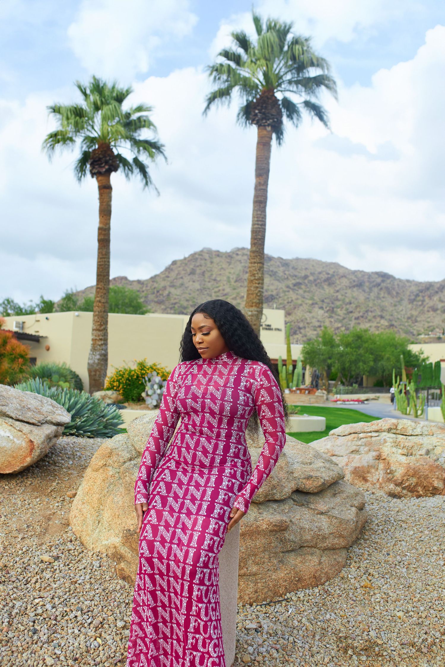 Person in pink patterned dress posing against desert landscape with palm trees and mountains.