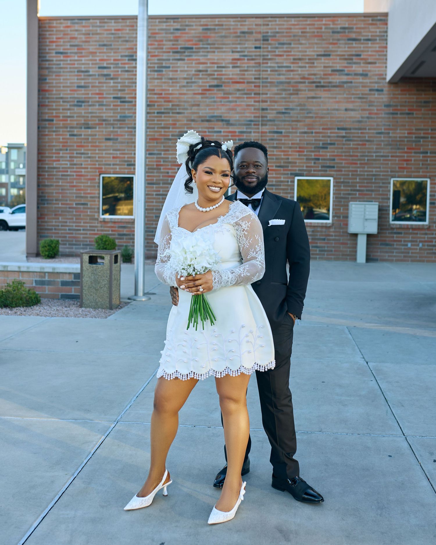 A couple in wedding attire poses together on a brick-lined sidewalk outside a building.
