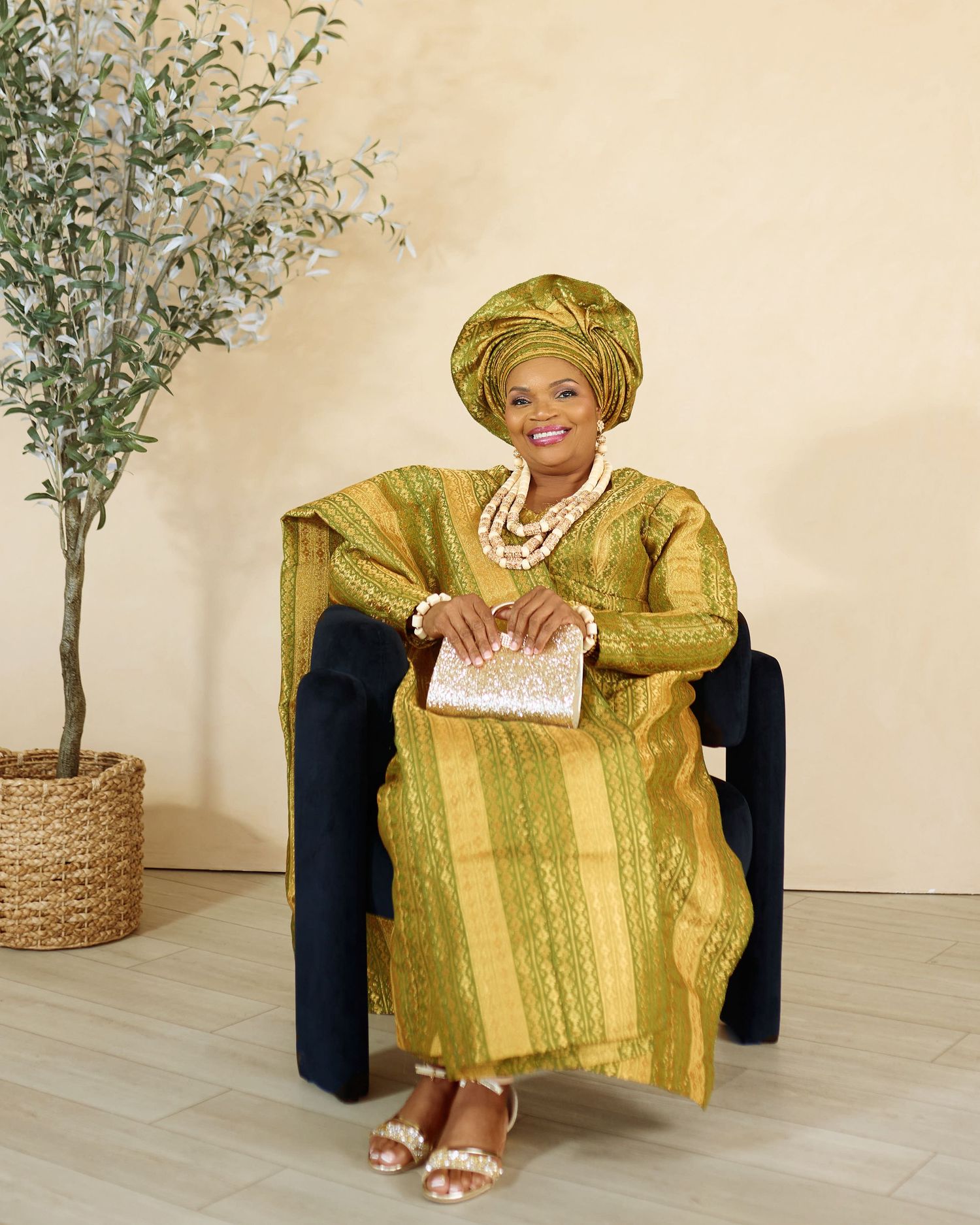 Person wearing traditional yellow African attire poses elegantly on chair with decorative plant.