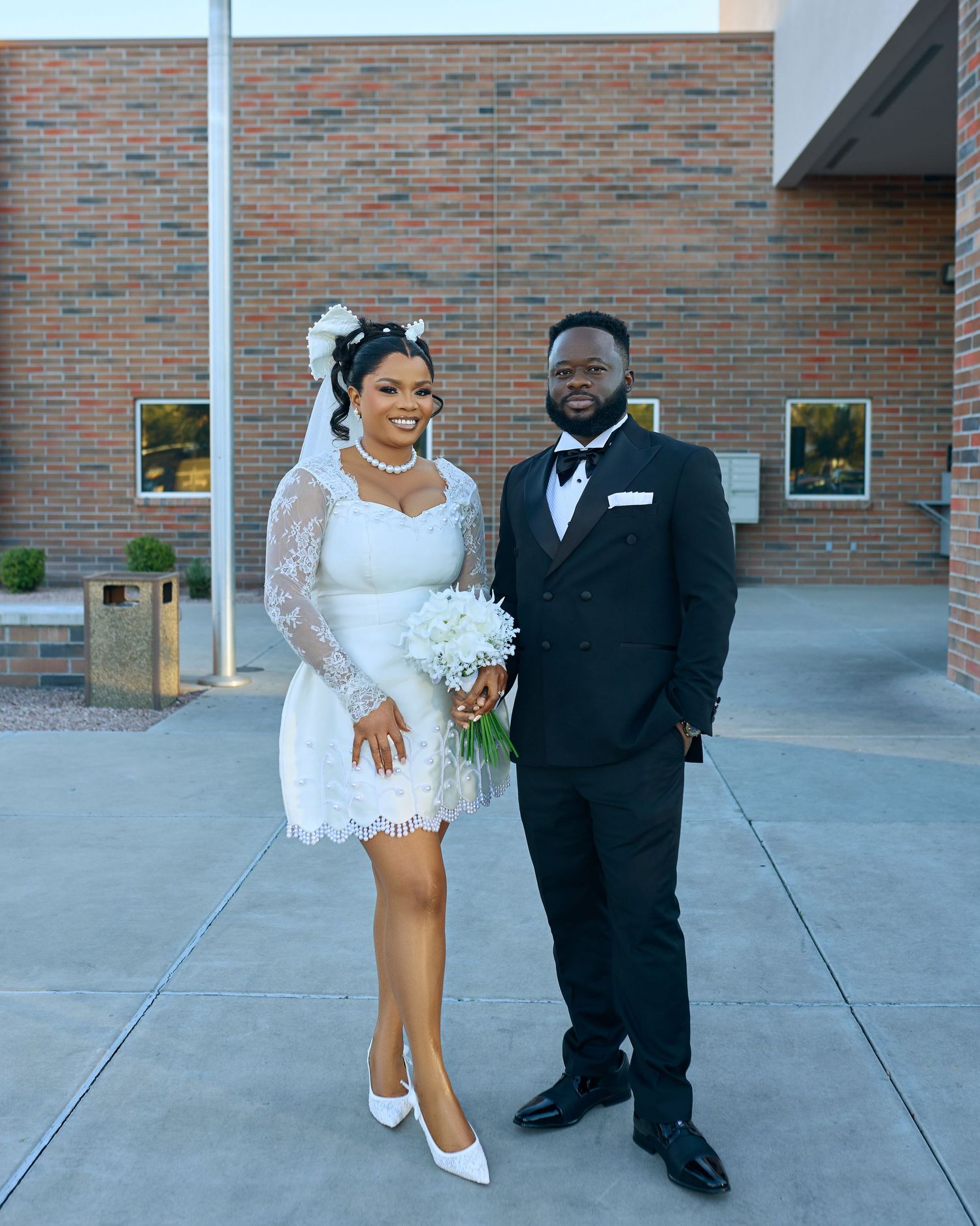 A couple in formal wedding attire poses together against a brick wall background.