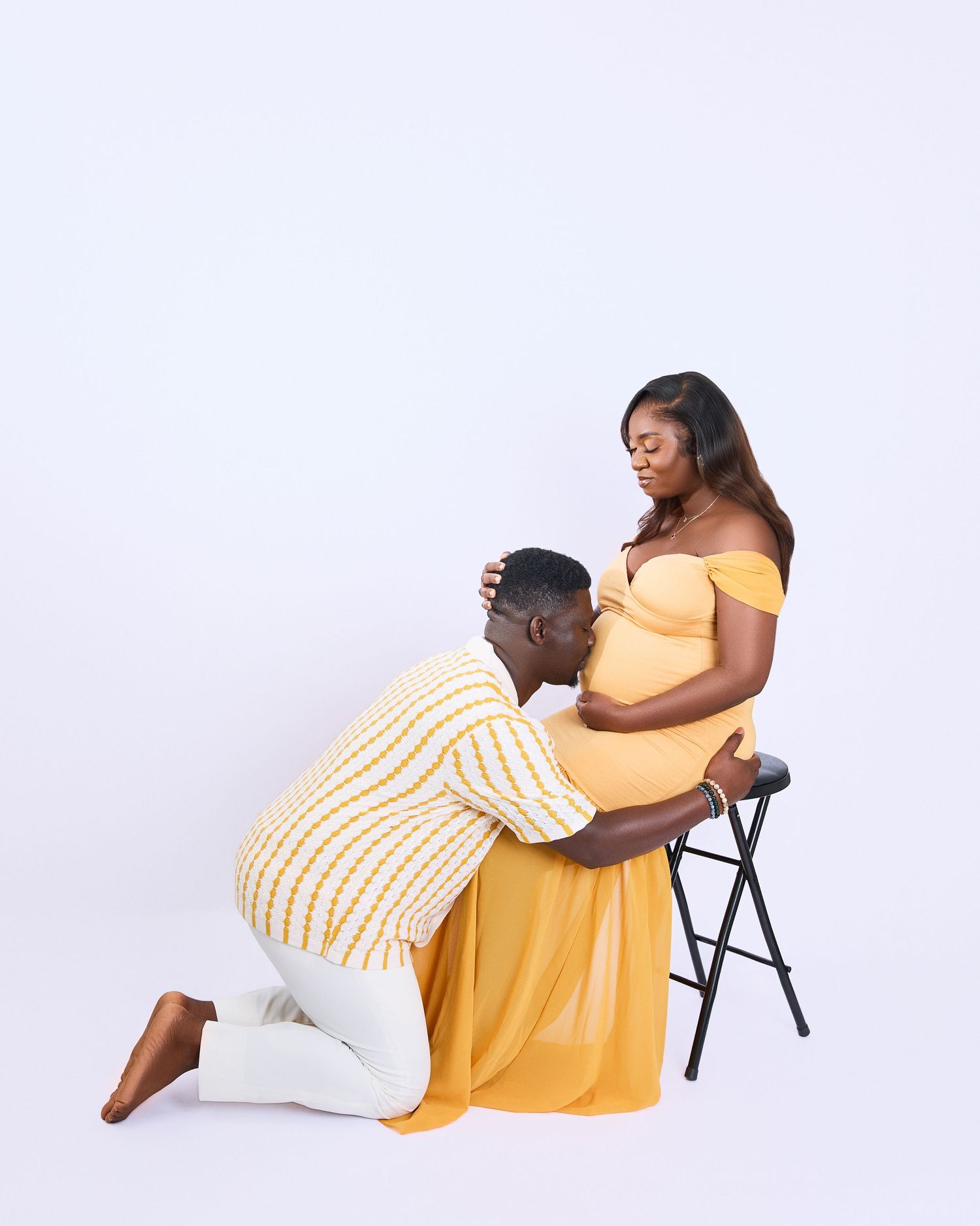 Intimate maternity photo with couple in yellow dress and striped shirt against white background.