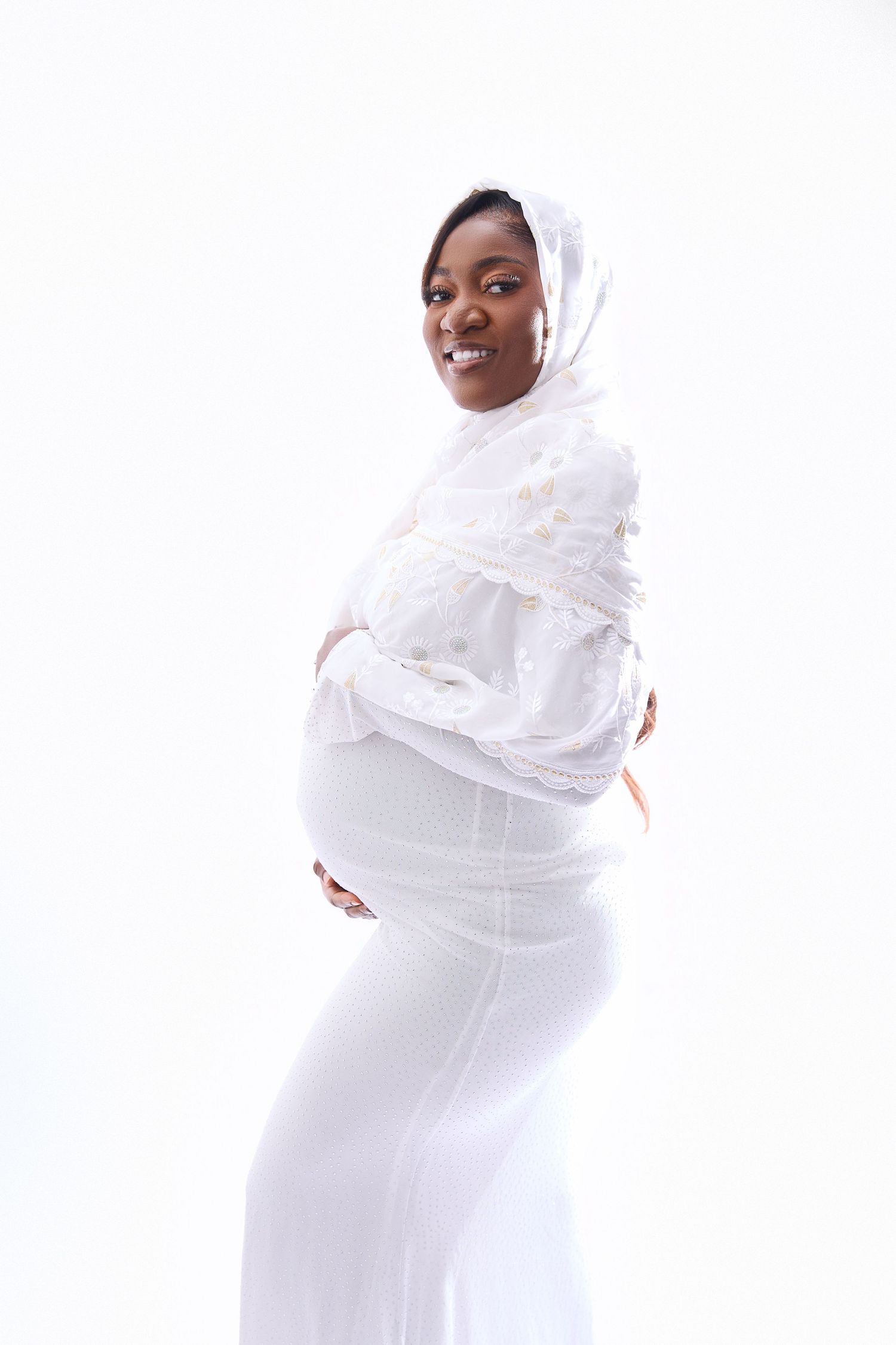 Maternity portrait featuring white flowing dress and head covering against light background.