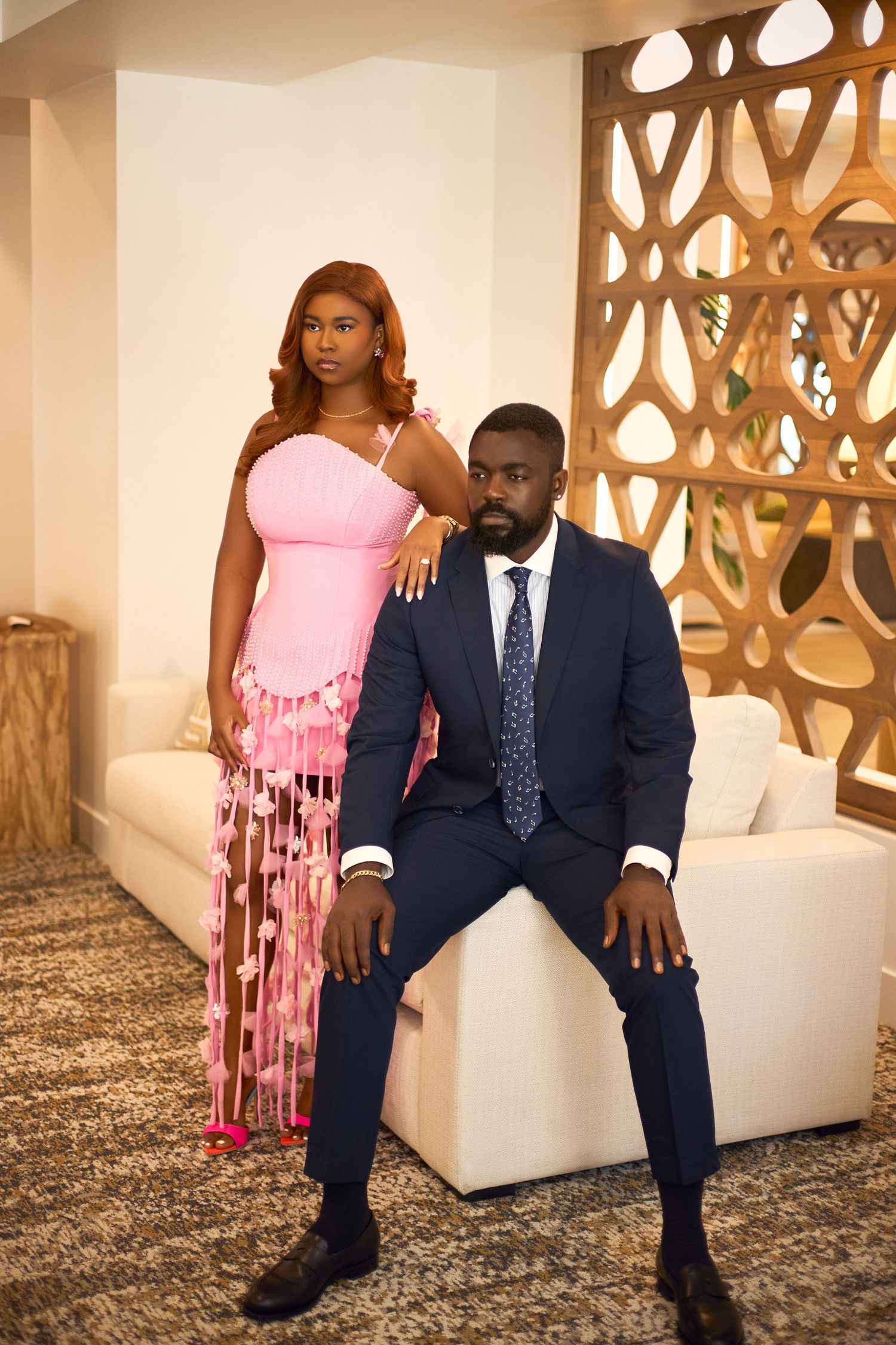Formal couple's portrait session with pink dress and navy suit against decorative wall backdrop.