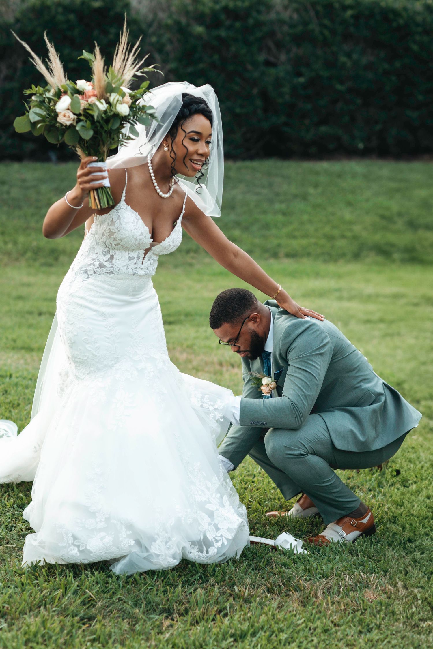 A couple in wedding attire playfully poses on grass during an outdoor photoshoot.