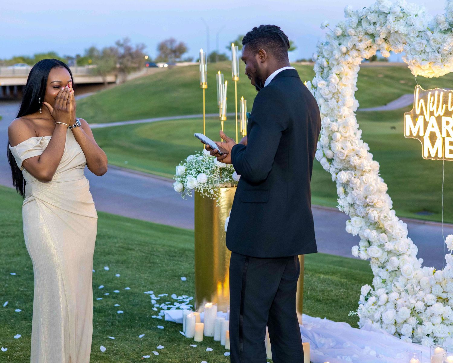Outdoor proposal scene with floral arch and 'Will You Marry Me' sign at sunset.