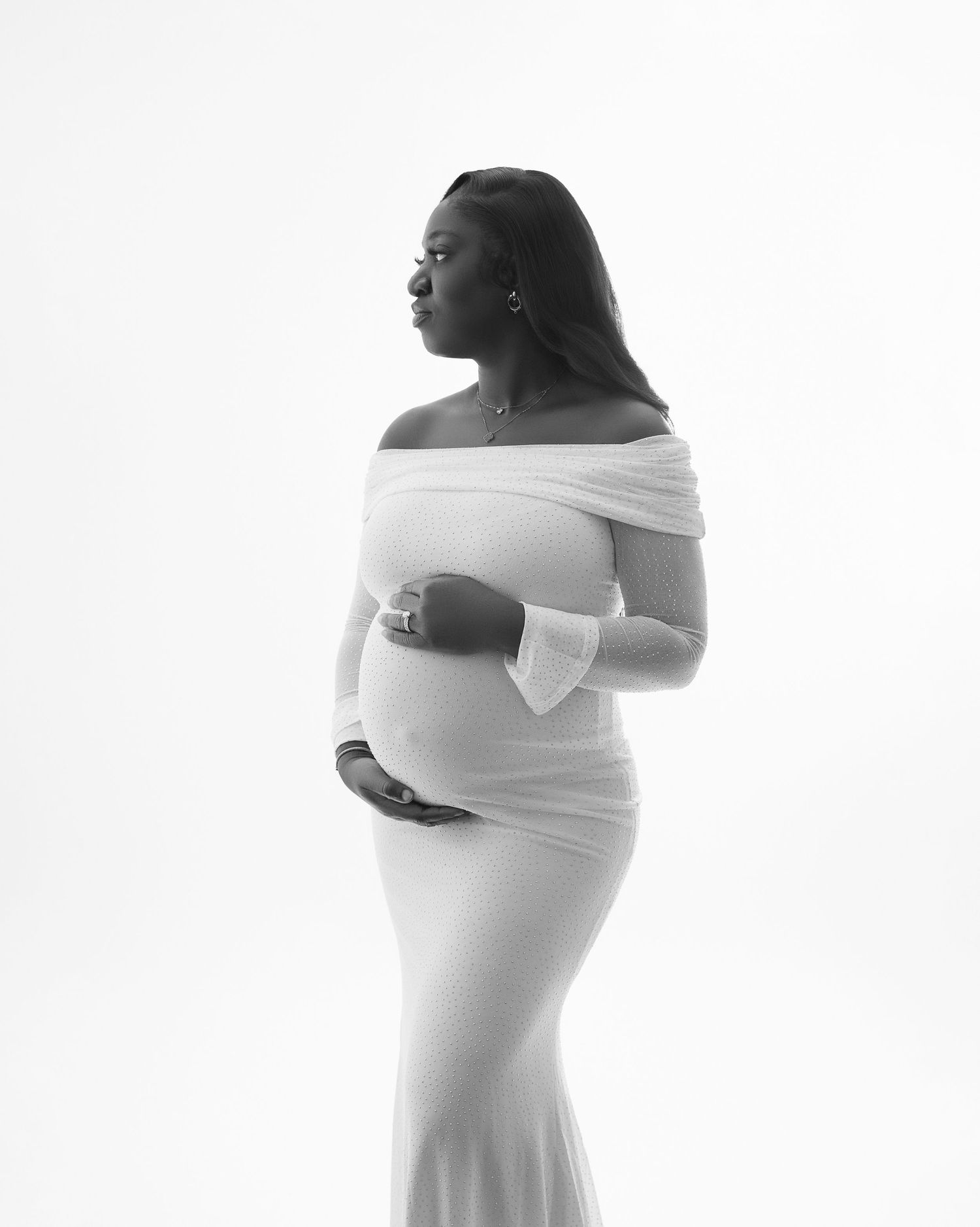 Artistic maternity photo in white off-shoulder dress against light background.