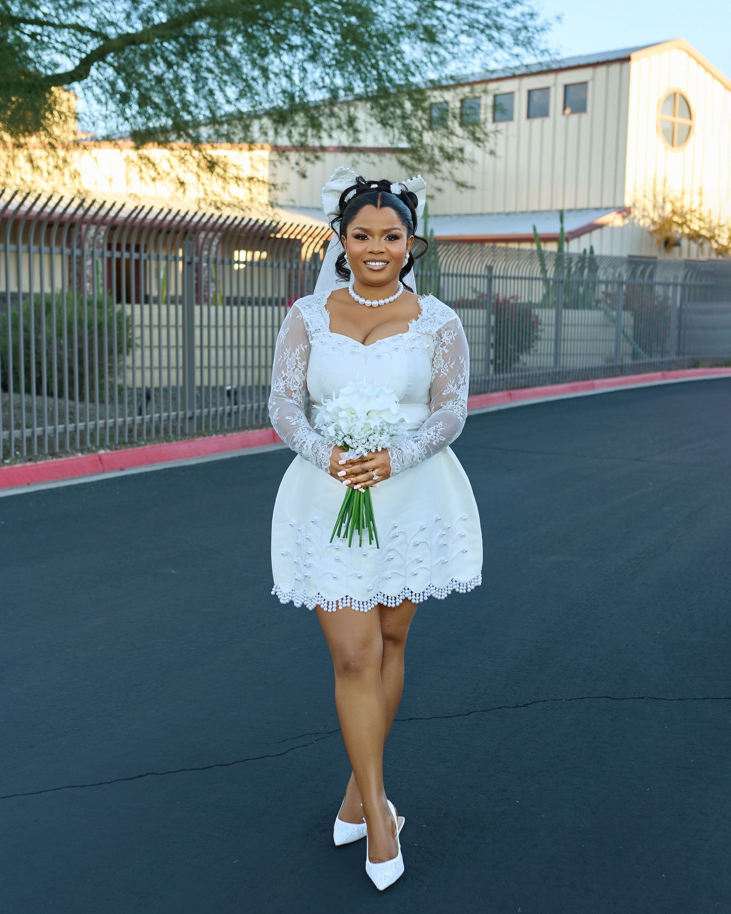 A bride in a short white lace dress and heels poses playfully outdoors on a sunny day.