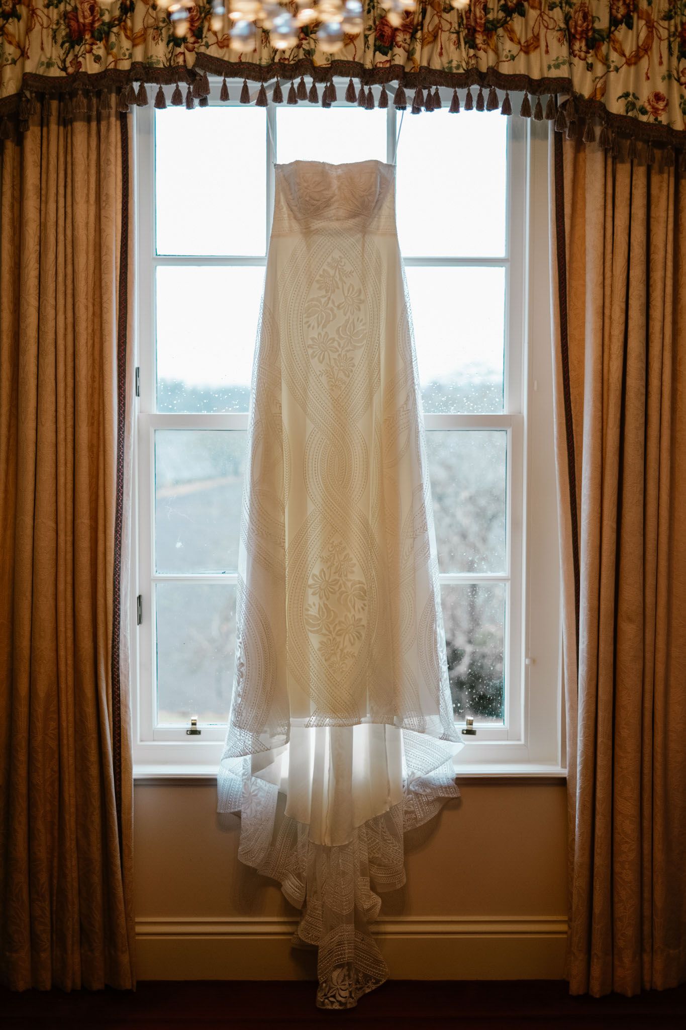 White lace wedding dress hanging in front of sunlit windows with golden curtains.