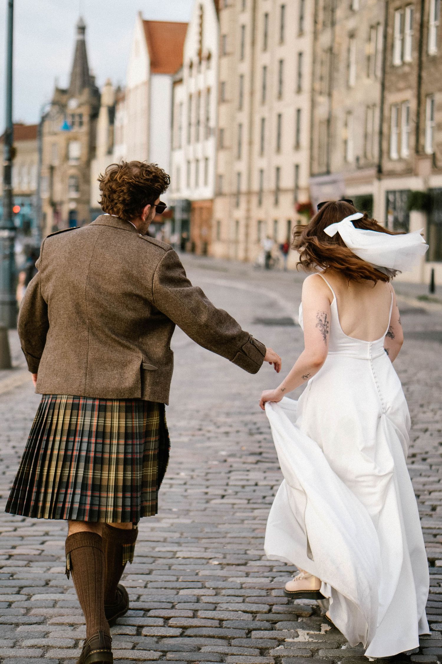 Couple runs along cobblestone street in Edinburgh with traditional Scottish attire and wedding dress.