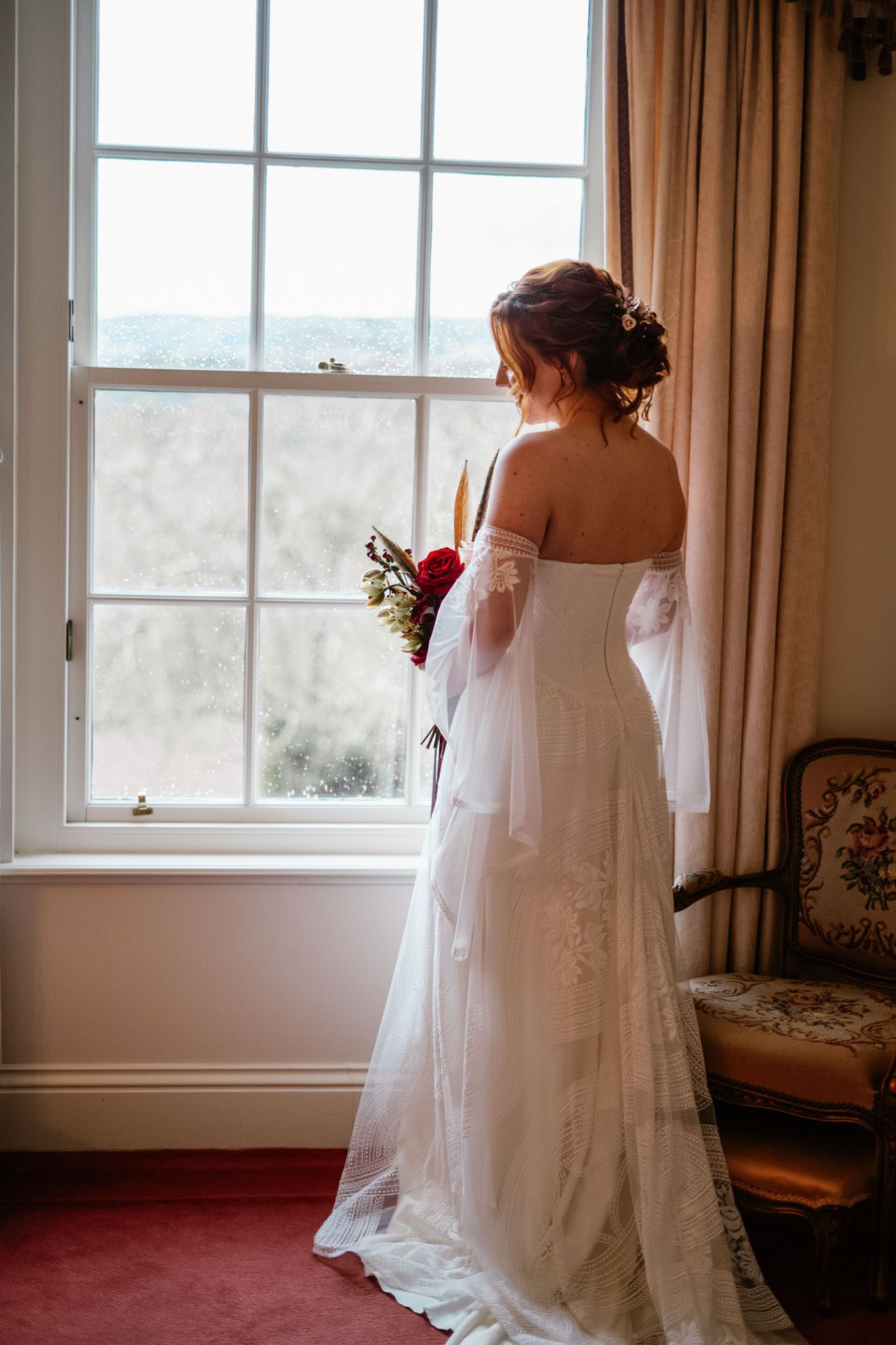 Bride in off-shoulder wedding gown standing by window with scenic view.