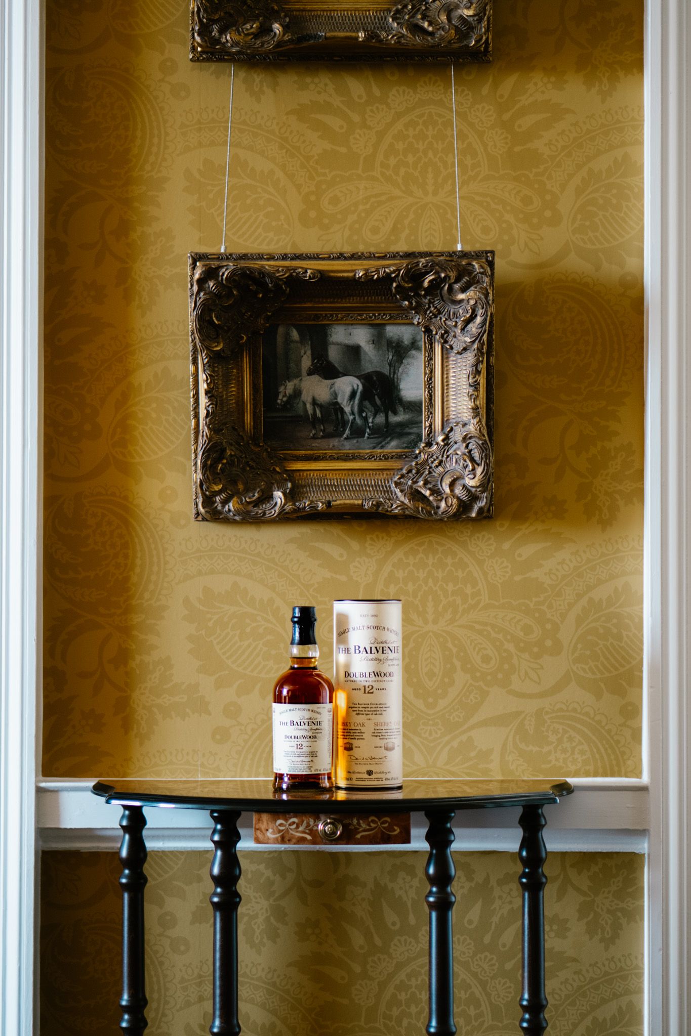Elegant interior with gold wallpaper and vintage frames displaying whiskey bottles on black tables.