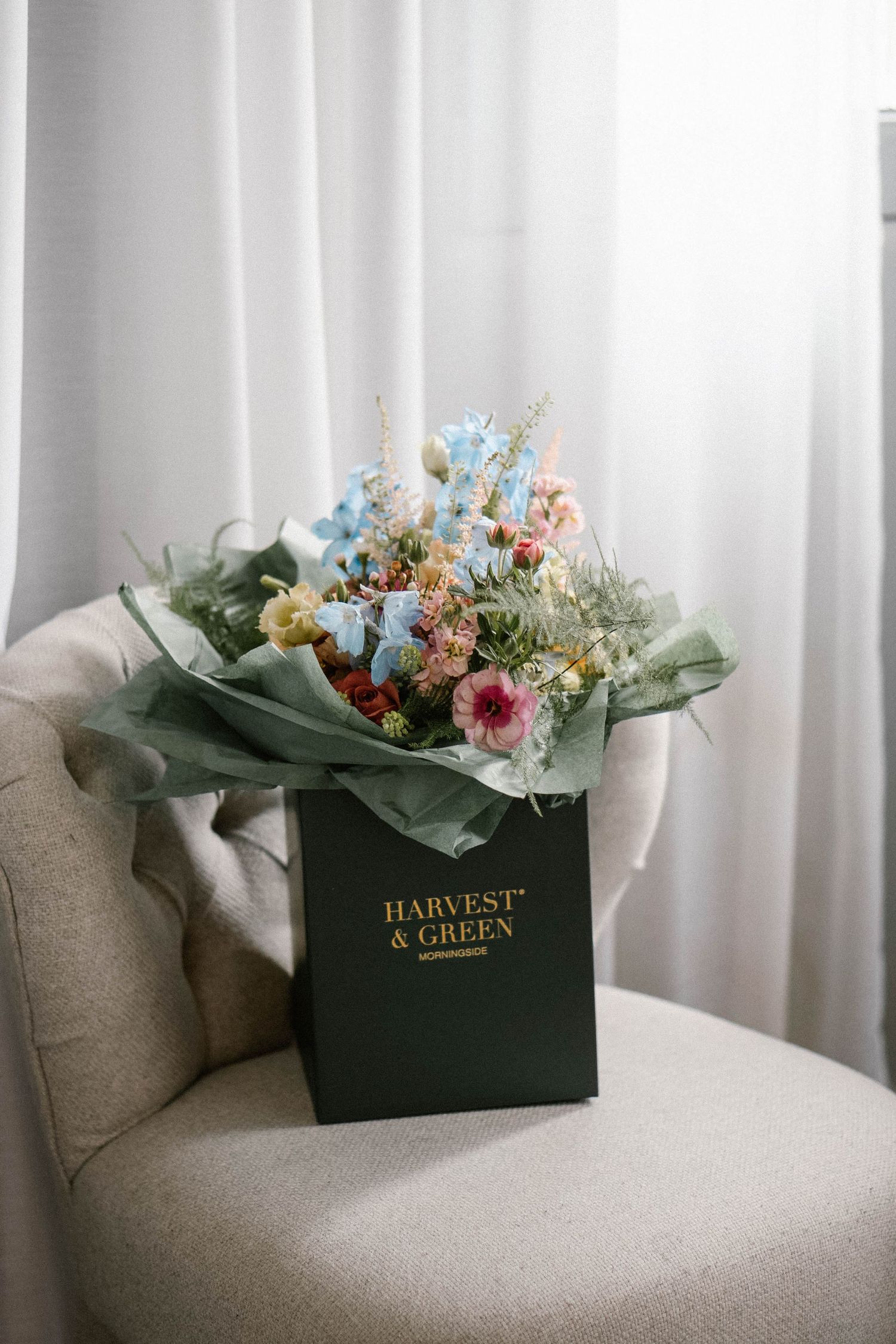 Floral arrangement in a green box sitting on a light gray couch against white curtains.