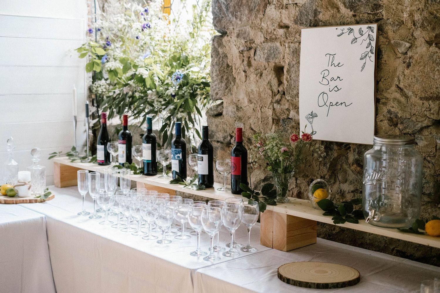 Wedding bar setup with wine bottles and glasses against a stone wall.