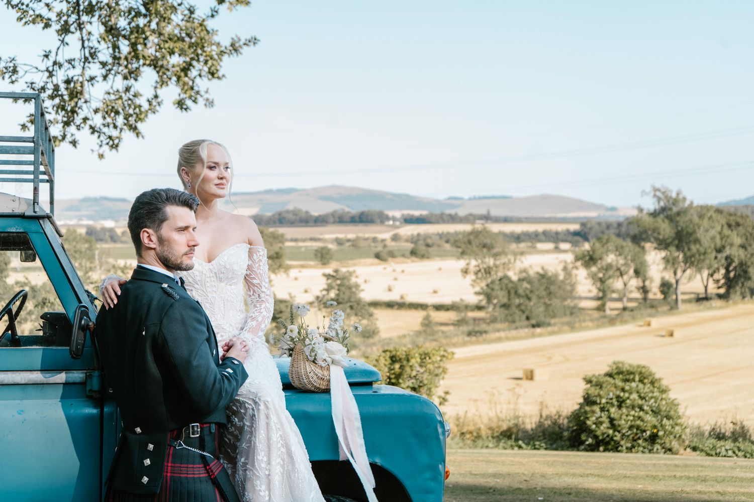 A couple stands together in an open field with rolling hills and trees in the background.