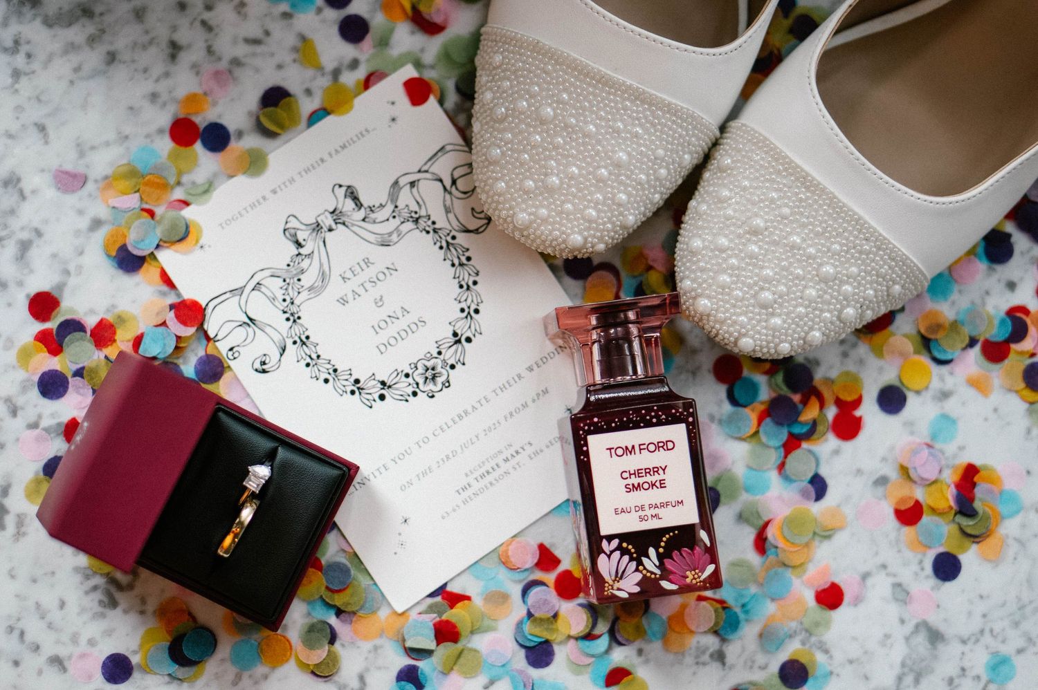 Wedding details flatlay with sparkly shoes, perfume bottle, and invitation on confetti-covered surface.