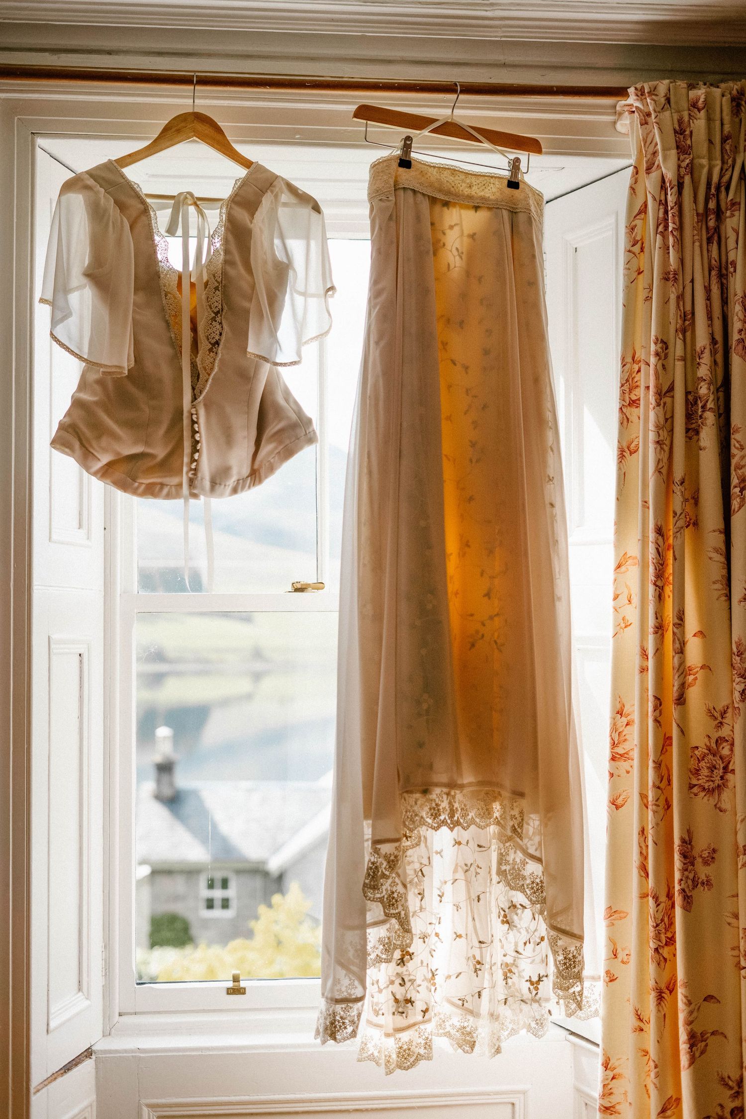 Wedding dresses and garments hanging in a sunlit window with floral curtains.