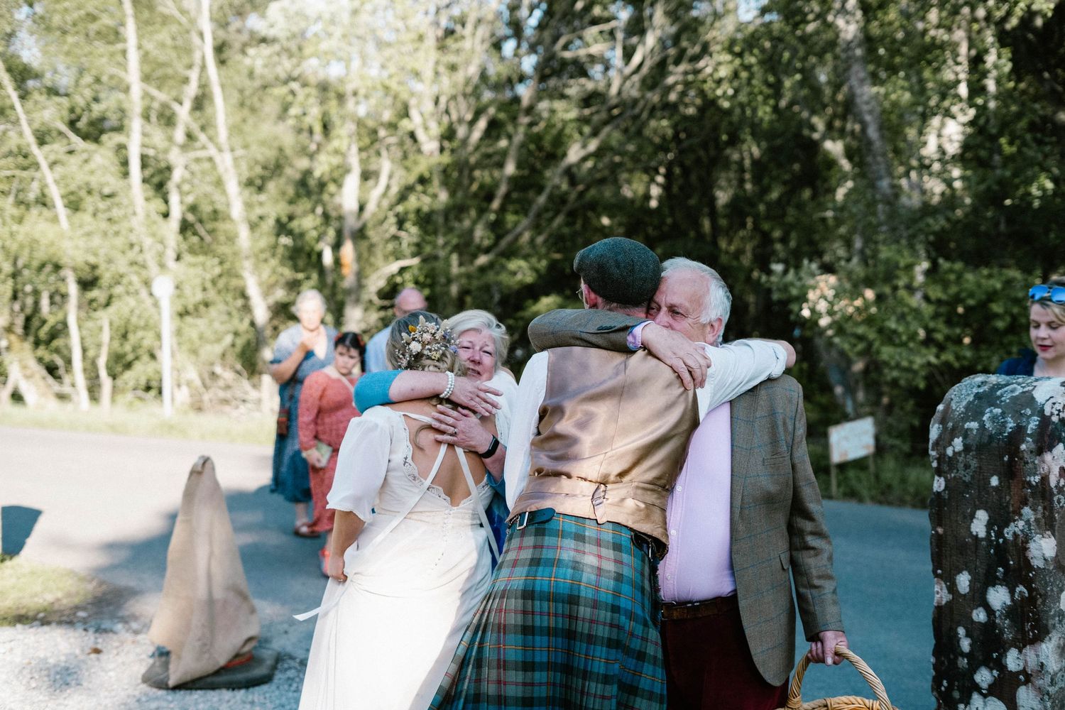 Outdoor wedding celebration with guests in traditional Scottish attire among trees.