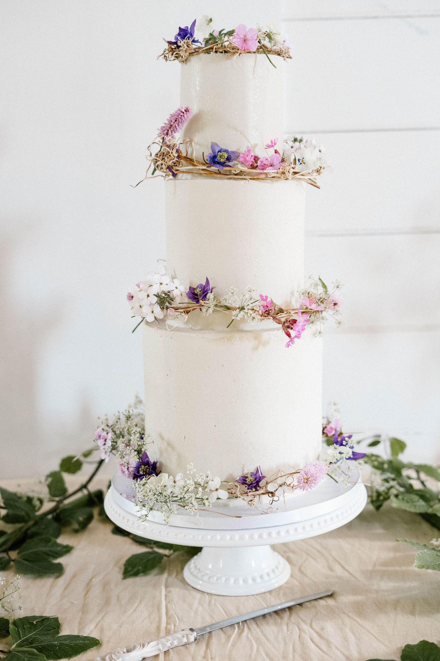 Elegant three-tiered white wedding cake decorated with delicate purple and pink wildflowers on a white cake stand.