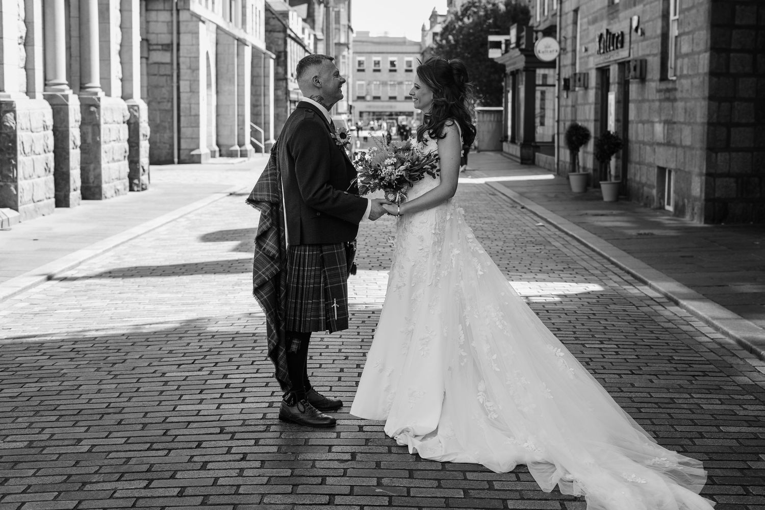 Black and white photo of a bride and groom in Scottish kilt exchanging vows on a cobblestone street.