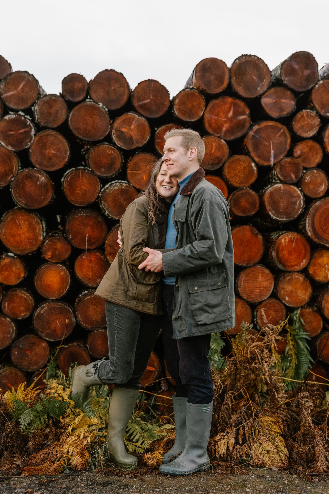 Couple in casual outdoor wear posing against stacked logs in a rustic setting.