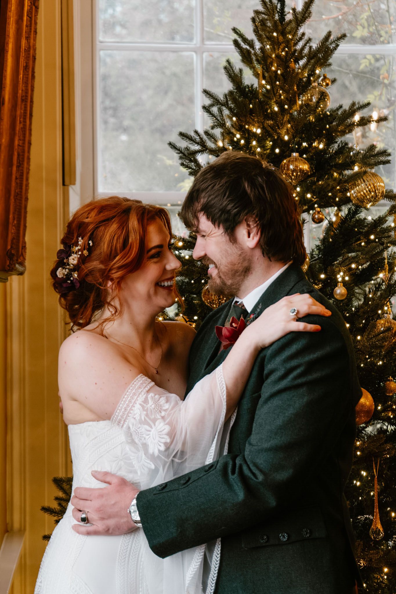 Wedding couple sharing intimate moments near decorated Christmas trees in sunlit room.