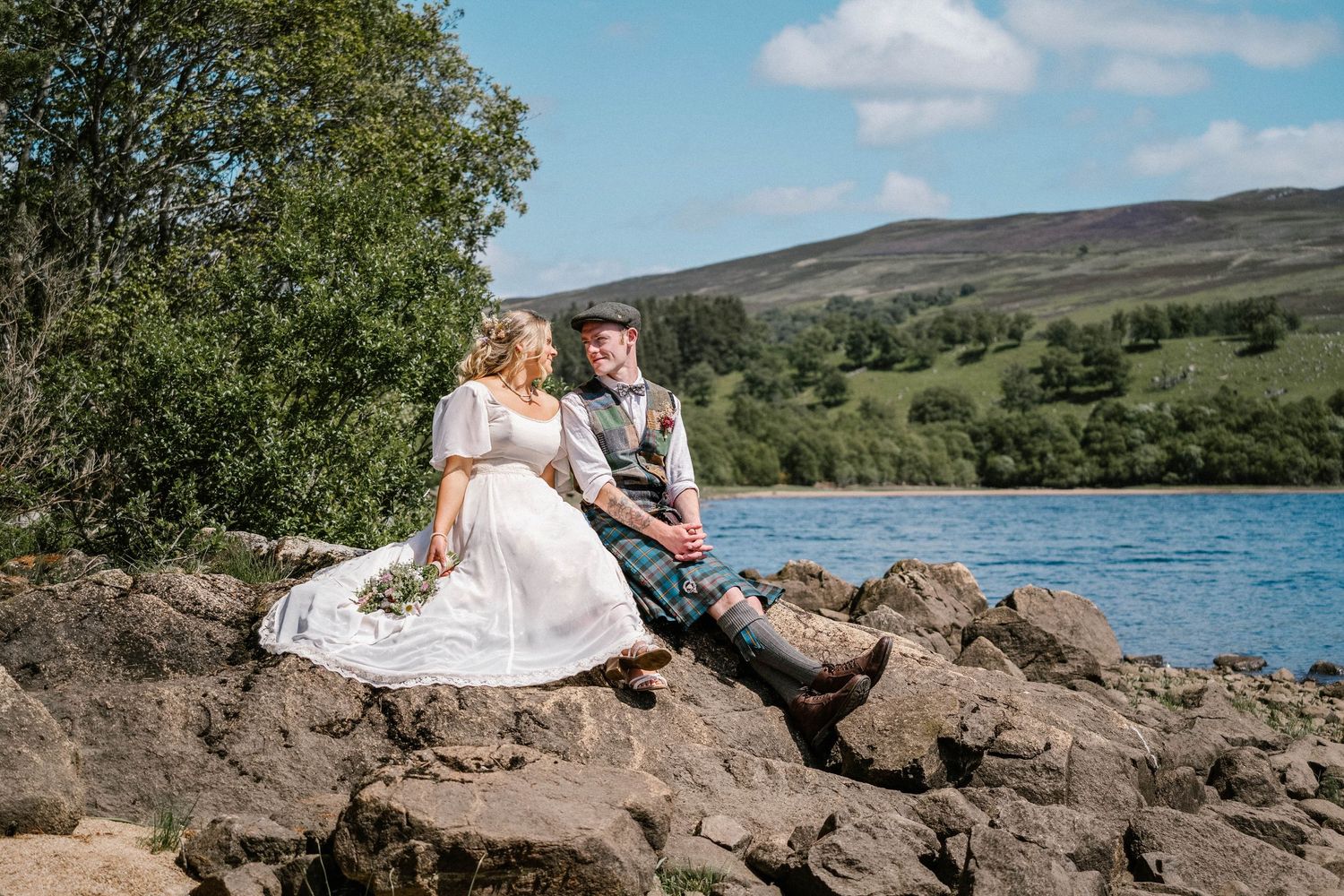 Newlyweds share an intimate moment on rocky coastline overlooking a serene lake.