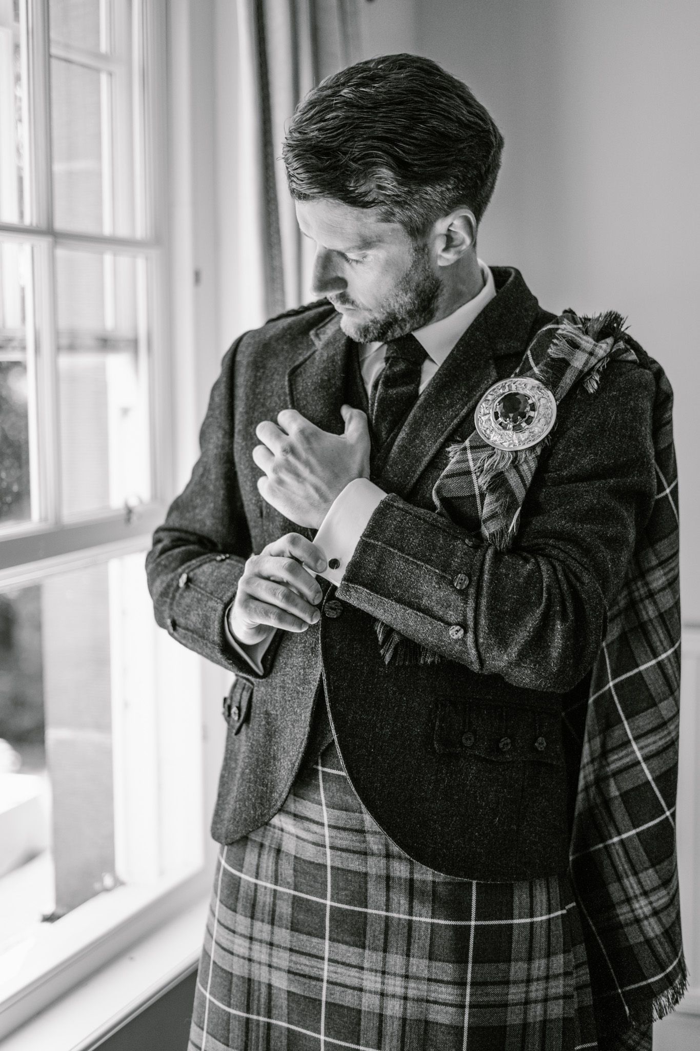 Man in traditional Scottish kilt and suit jacket wiping tears by sunlit window.