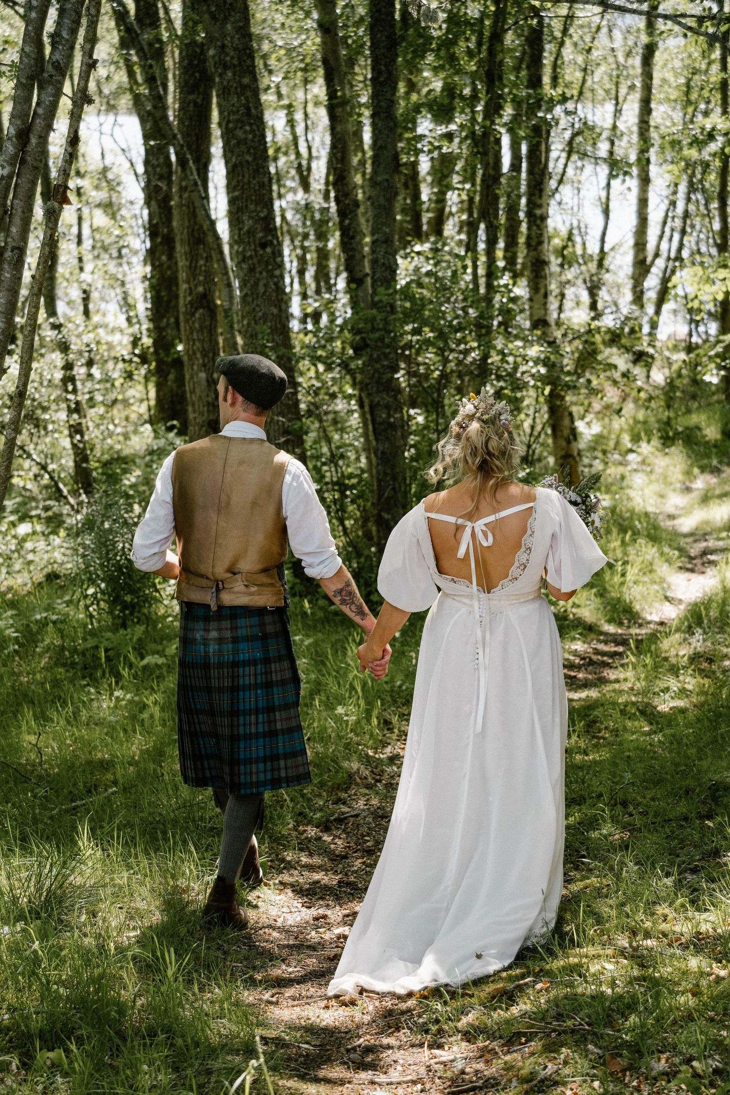 A couple walks hand in hand through a sunlit forest path wearing traditional Scottish wedding attire.