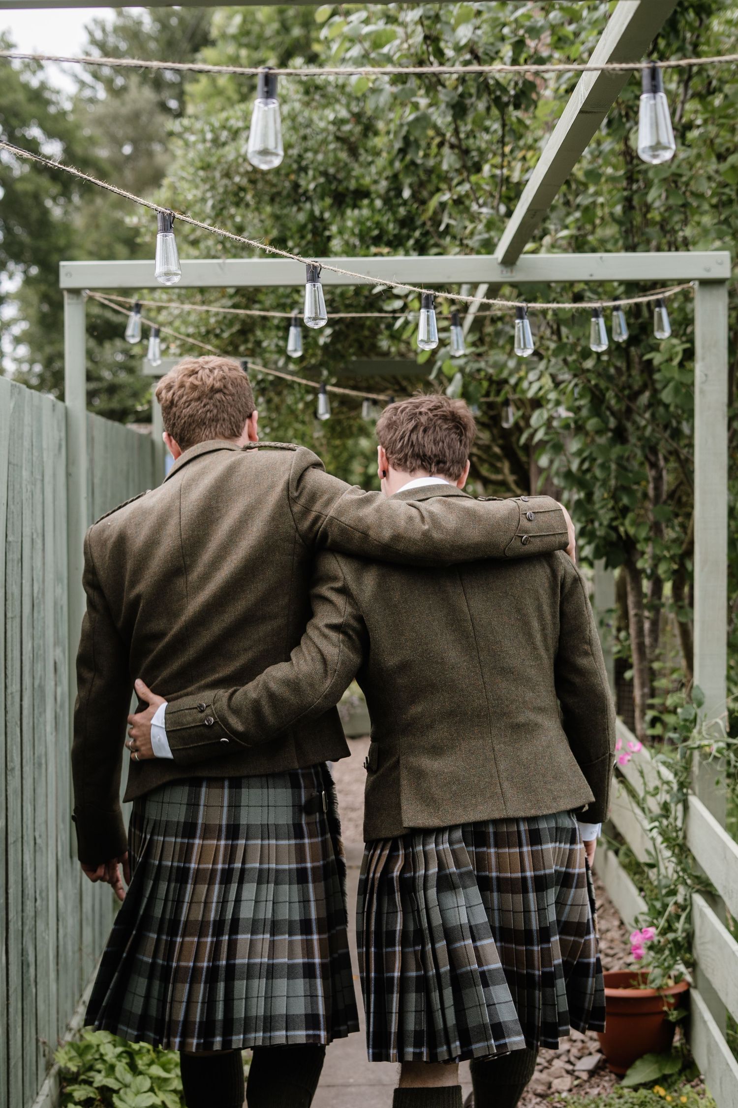 Two men in matching tartan kilts and tweed jackets walk arm in arm through a garden decorated with string lights.