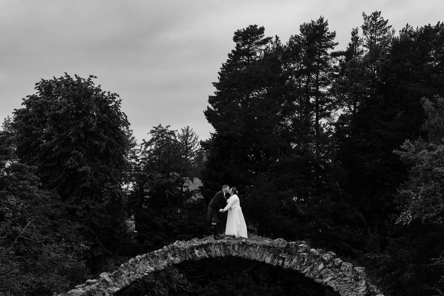 Black and white photo of a couple kissing on a stone arch bridge surrounded by lush trees.
