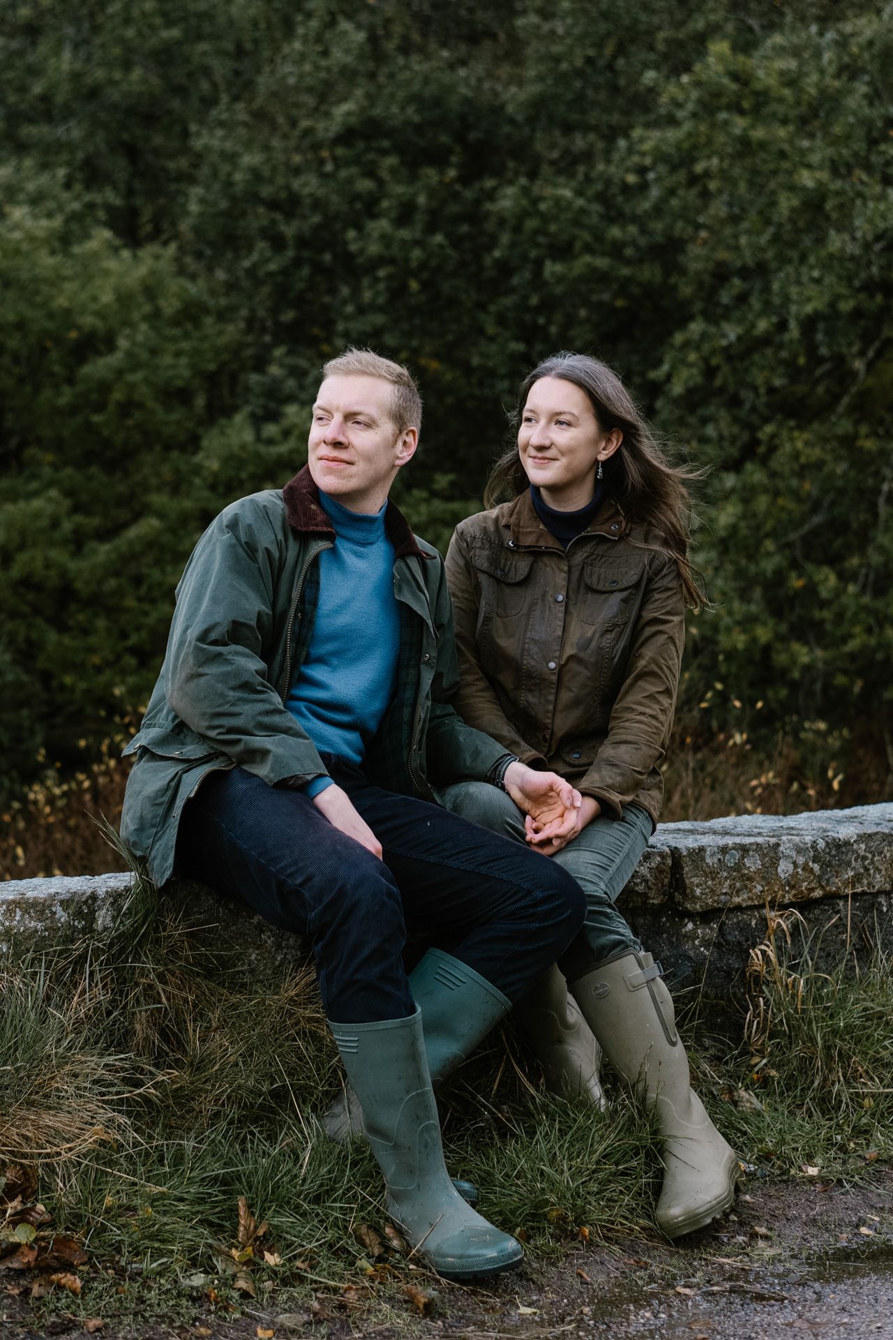 Two people sitting on a stone wall surrounded by lush greenery on an autumn day.
