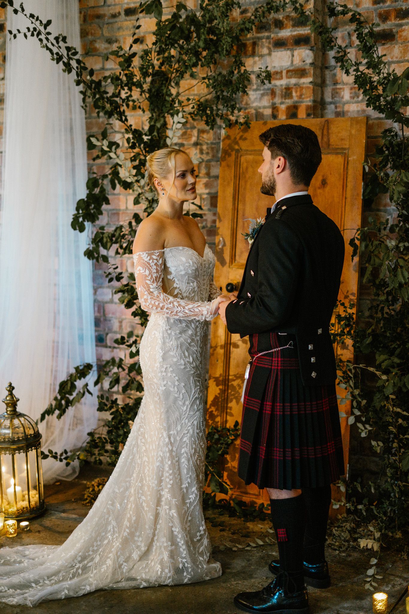 A romantic wedding scene with a white lace dress and traditional Scottish kilt against a rustic brick wall with ivy.