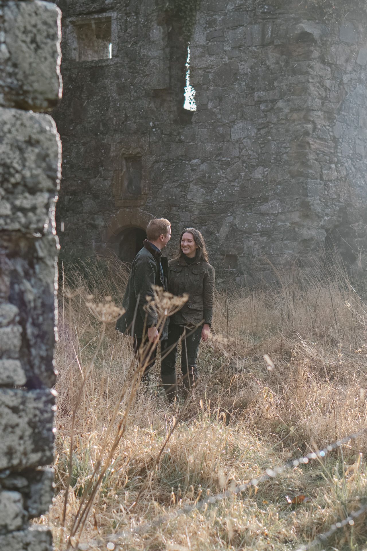 A couple walking through tall grass near ancient stone ruins on an overcast day.